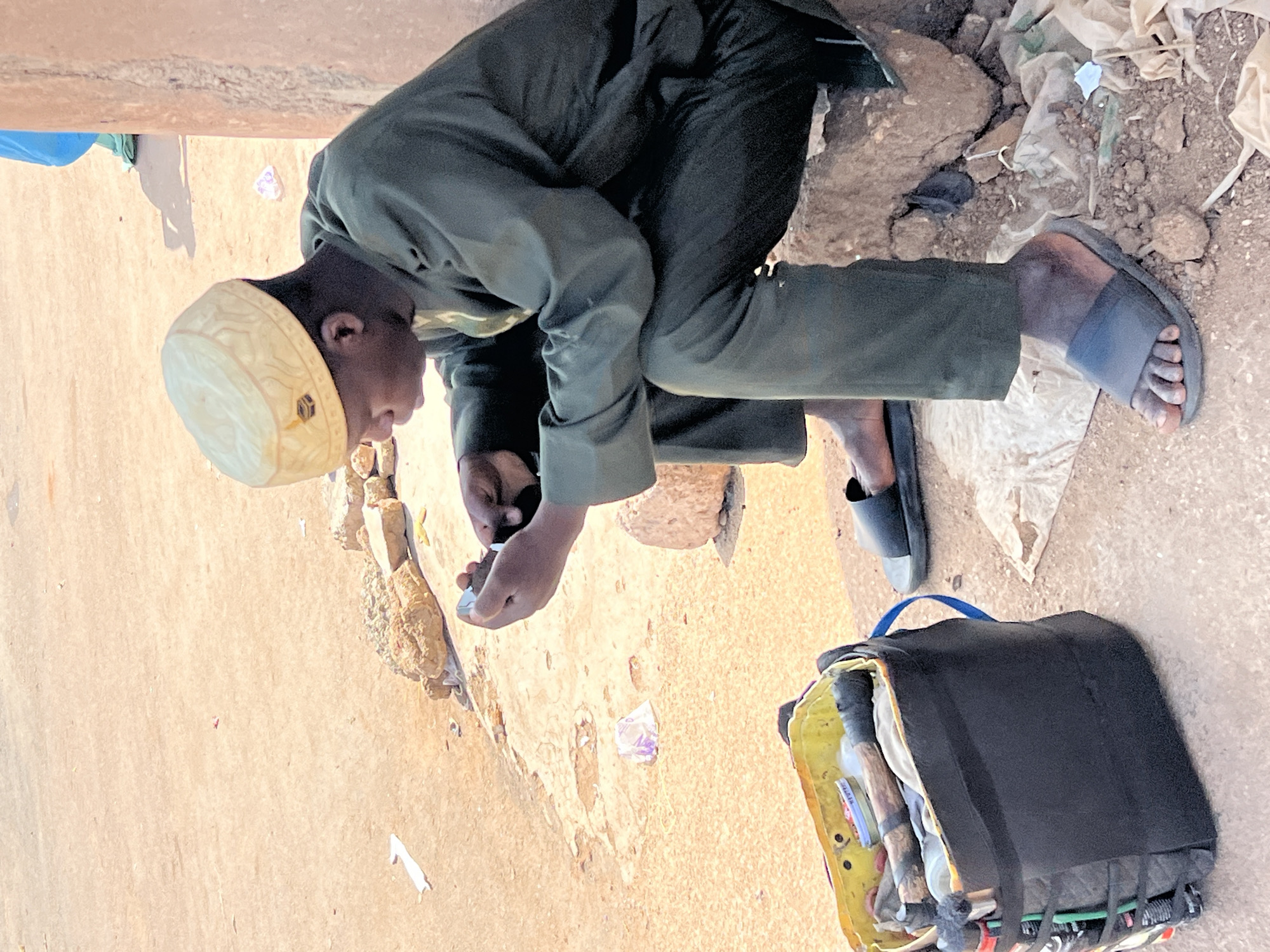 A young man wearing a white kufi cap and dark clothing kneels on the ground in an outdoor setting. He appears to be examining or working with a small object in his hands. Next to him is an open bag with various items inside, possibly merchandise or supplies. The ground appears to be rough and sandy, and there is debris, including some plastic or paper waste. The scene suggests an urban environment, possibly a marketplace or street corner, in Jos, Nigeria.