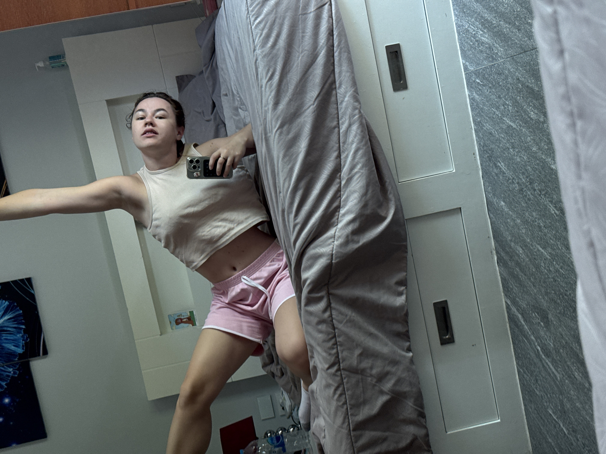 A person is lying on a bed in a room, taking a selfie with their phone. The person is wearing a light-colored tank top and pink shorts. They are leaning back on their left arm, with their right arm extended upwards. The bed has a grey comforter with a subtle pattern. In the background, there is a white headboard with a picture frame on it, and a bottle of water on a bedside table. The room appears to be a bedroom, possibly in a hotel or residential setting. The lighting suggests it is daytime, with natural light coming from a window or the overhead room lighting. The image does not provide any specific visual cues to indicate the location is Rawai, Thailand.