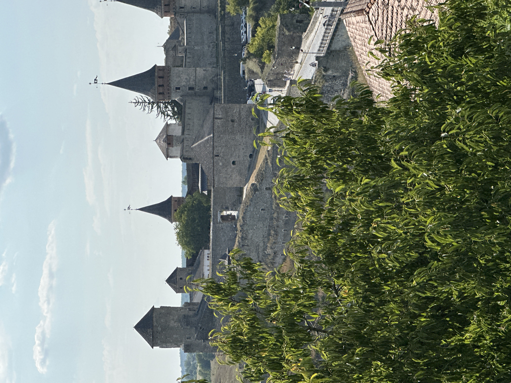 The image depicts a panoramic view of the Kamianets-Podilskyi Castle in Ukraine, a prominent historical fortress. In the foreground, lush green foliage partially obscures the view of the castle complex, which is constructed from grey stone. Several towers with conical roofs are visible, some topped with weather vanes. The castle walls are extensive, with various architectural elements and openings. A road winds its way towards the castle, with a bridge and several people walking on it. Vehicles are parked in a lot near the castle entrance. The sky above is a clear blue with scattered white clouds. The overall scene conveys a sense of history and grandeur, with the natural elements of the foliage and sky complementing the imposing stone architecture.