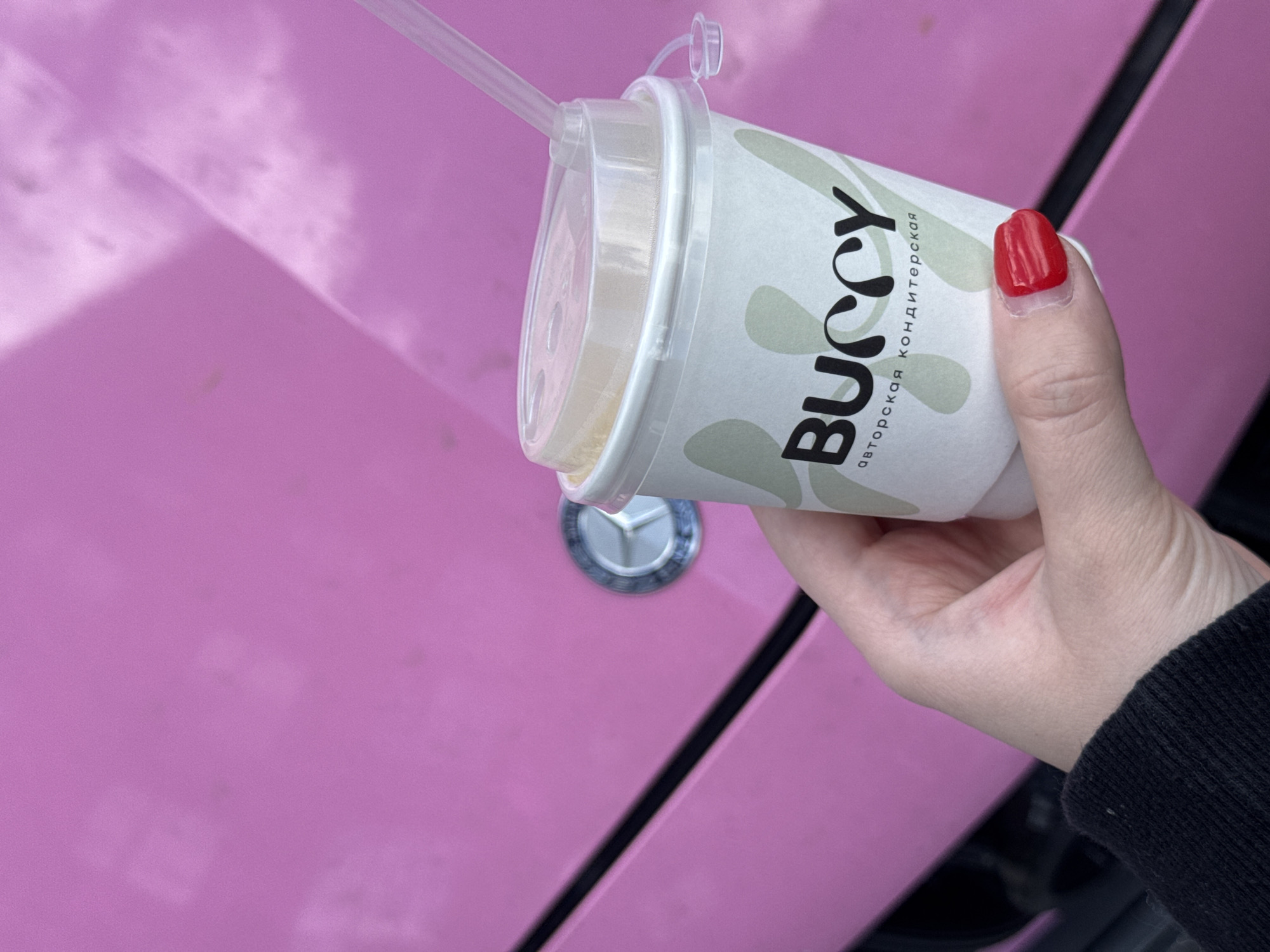 A hand with a red-painted nail holds a paper cup with a plastic lid and straw. The cup has a white background with a green design and black text. The text on the cup reads "BUCCY" in large letters, and below it, in smaller letters, "авторская кондитерская" (which translates to "author's confectionery"). The cup is tilted towards the viewer, showing the straw and the top of the drink.
The background is a bright pink surface, which appears to be the hood of a car, with a circular emblem visible on it. The emblem is silver and circular, with three wings radiating from the center, strongly resembling a Mercedes-Benz logo. The lighting suggests it is daytime, with natural light illuminating the scene. The overall impression is that someone is holding a beverage from a confectionery while leaning against or standing near a pink car. The scene appears to be outdoors, given the lighting.