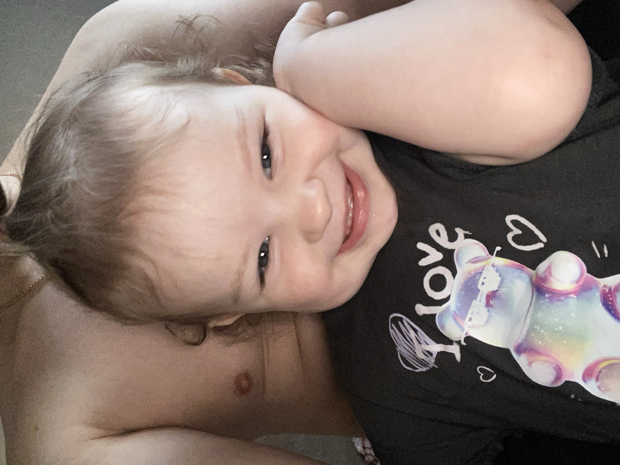 A close-up, high-angle shot captures a joyful young child, likely a toddler or baby, lying on their back and looking directly at the camera. The child's head is tilted to the right, resting on what appears to be an adult's fair-skinned arm or chest, parts of which are visible in the foreground and background, including a nipple and some hair.
The child has light-colored, somewhat curly hair and fair skin, with bright, alert eyes and a wide, open-mouthed smile, showing visible teeth. Their expression conveys happiness and contentment.
They are wearing a black t-shirt that features a prominent, colorful, iridescent gummy bear design. To the left of the gummy bear, the word "love" is written vertically in white, accompanied by a small white heart. Another abstract white drawing, possibly a wing, is also on the shirt. The background is out of focus, suggesting an indoor setting, possibly a bed or couch. The lighting is soft and even, highlighting the child's features.