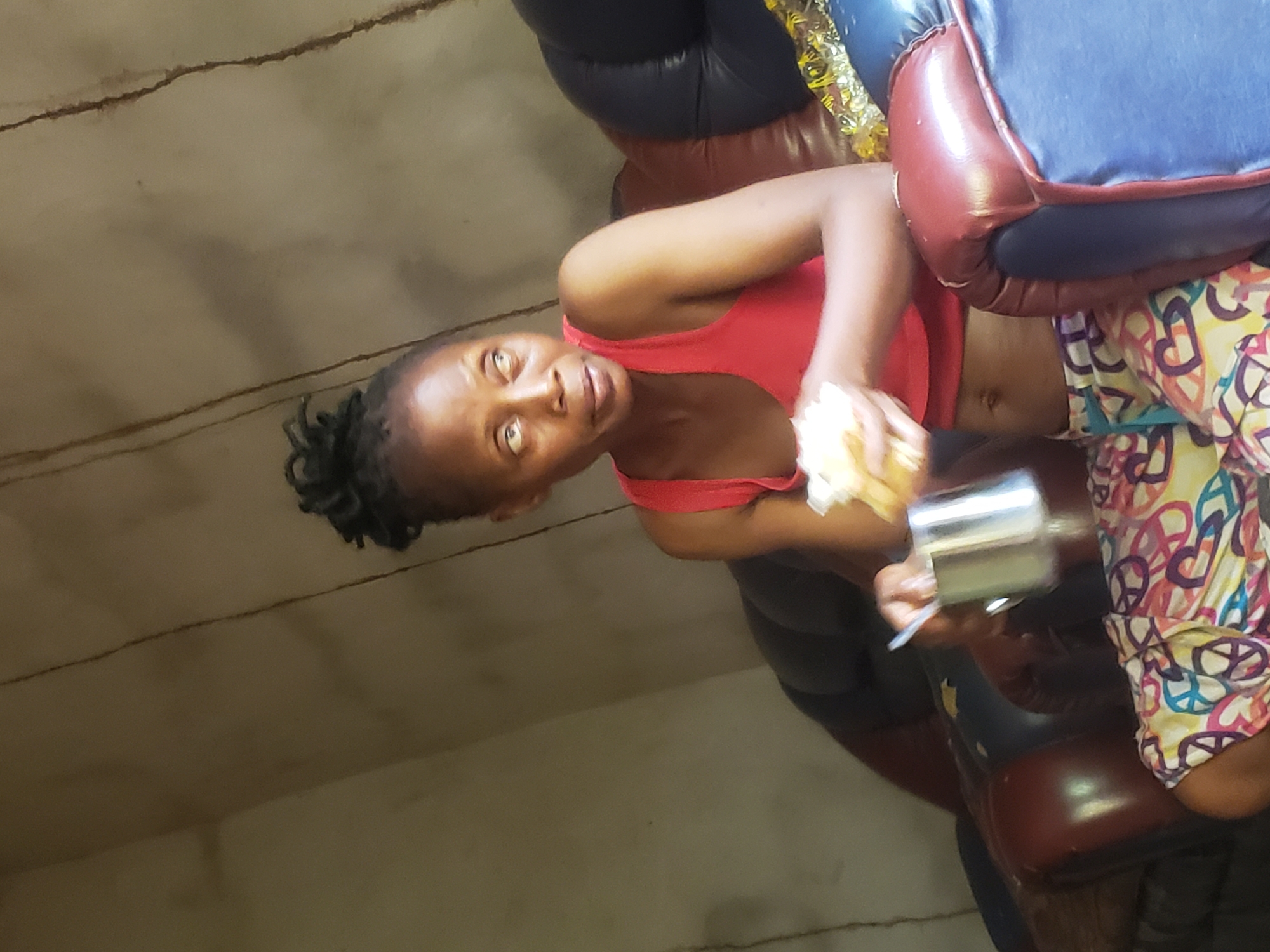 An indoor shot shows a young Black woman seated, looking directly at the camera with a calm expression. She has dark hair styled in small braids or twists. She is wearing a red tank top and a colorful garment on her lower half, patterned with hearts and peace signs in yellow, pink, purple, and blue. In her right hand, she holds a light-colored, textured item, possibly food. Her left hand holds a metallic cup or container and a thin white stick or spoon. She is positioned on a multi-colored sofa or chair with brown leather-like sections and a blue cushion. The background consists of a light brown or beige wall with several distinct horizontal dark lines or cracks, and some shadows are also visible on the wall. The lighting suggests an indoor setting. No text is visible.