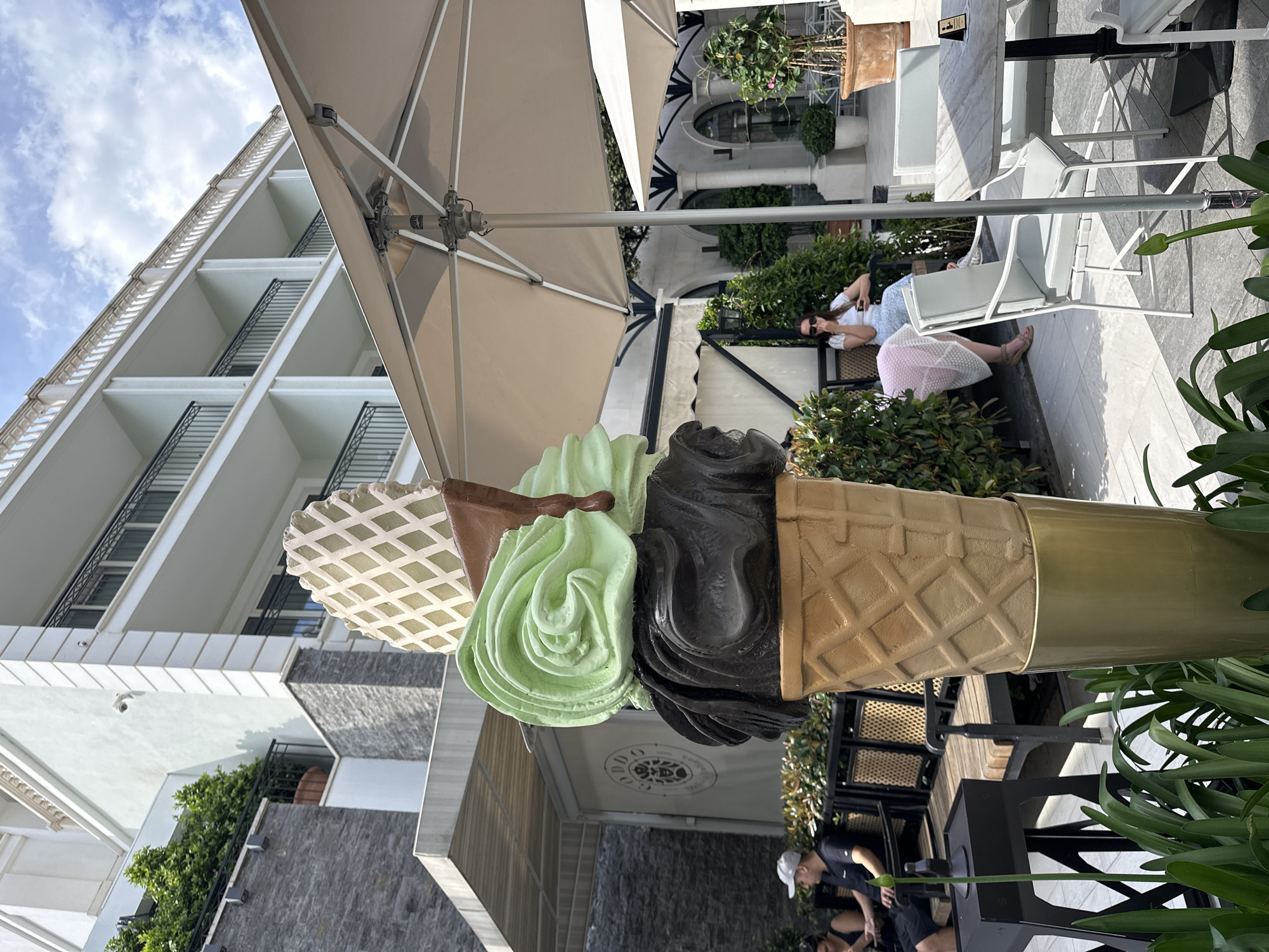 The image shows an outdoor cafe setting in Budva, Montenegro. In the foreground stands a large decorative ice cream cone sculpture with three scoops: white, green, and black. The cone's base is covered in a waffle pattern and is attached to a pole.
Behind the sculpture, several people are present. A woman lounges on a white chair under a large beige umbrella, while other people are seated at nearby tables. The background includes a multi-story building with white balconies and black railings. Green plants and shrubs add to the cafe's ambiance. The sky is partly cloudy.