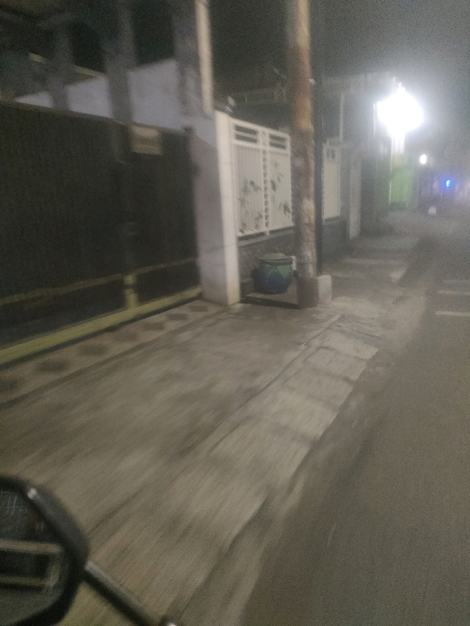 The image depicts a street scene at night in Surabaya, Indonesia. The perspective appears to be from a moving vehicle, possibly a motorcycle, as indicated by the blurred handlebars in the lower left corner. The street is paved and appears damp, suggesting recent rain or a humid environment.
On the left side of the street, there is a dark, closed gate, possibly leading into a property or business. Beyond the gate, a building with a white facade is visible. Next to the gate, a white metal fence with a decorative pattern separates the walkway from the property. In front of this fence, there is a large, dark green cylindrical object, which could be a trash bin or a decorative planter.
A wooden pole, likely a utility pole, stands prominently in the middle ground, casting a shadow on the sidewalk. The sidewalk itself is made of concrete and shows signs of wear and tear, with some darker patches possibly indicating stains or moisture. Further down the street, a bright light source illuminates the scene, creating a hazy glow that obscures details in the distance. There are other faint lights visible, one appearing blue, suggesting traffic or streetlights. The overall atmosphere is hazy and somewhat indistinct due to the low light conditions and motion blur. The architecture visible is typical of urban areas in Indonesia, with a focus on functional design.