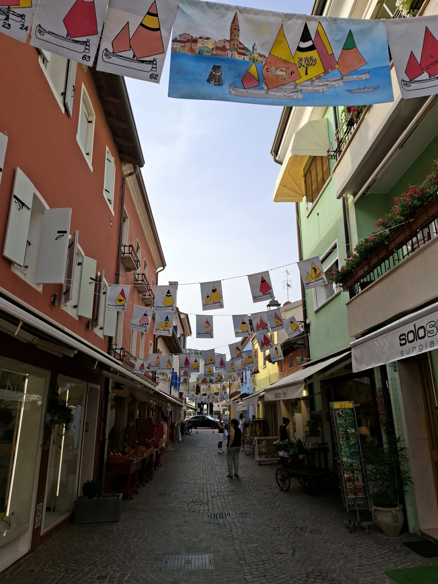 This is a medium-shot, eye-level, outdoor, daytime photograph taken in the city of Caorle, Italy, on a cobblestone street lined with buildings and shops. The street is decorated with a string of triangular flags, many of which feature simple drawings of sailboats.
On the left, a two-story building with a reddish-orange facade has multiple windows with white shutters, some of which are open. A shop with large glass windows and bright interior lighting is visible at the street level.
On the right, a two-story building with a light green facade features balconies with planters overflowing with pink flowers. Above the balconies, awnings extend over windows. A shop called "solo sunglasses" is visible at street level.
Several people are present in the scene. Two women are walking in the distance, and another person stands near a display rack of items. A dark-colored car is parked further down the street. The sky above is clear and pale blue. The overall impression is that of a picturesque European street scene during a summer day.