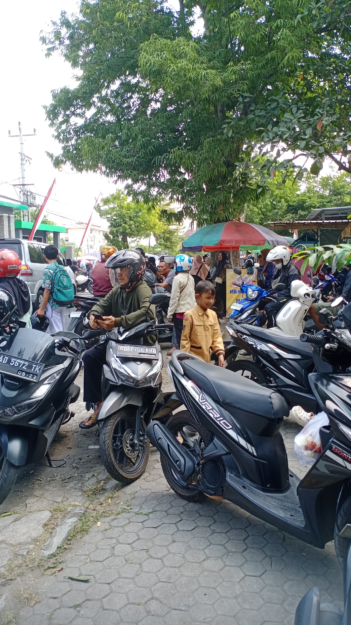 The image depicts an outdoor scene in Joho, Indonesia, likely a parking area or street lined with trees. In the foreground, several motorcycles are parked. A man wearing a dark green long-sleeved shirt, dark pants, and a grey helmet with a clear visor is seated on a silver motorcycle, holding a smartphone. To his left, another motorcycle is parked, with a license plate reading "AD 5672 TK 05-27". To the right of the man, a young boy in a tan uniform stands. Further back, more people and motorcycles are visible, some under colorful umbrellas. A silver car is parked on the left side of the image. The ground is paved with hexagonal tiles. Lush green trees with dense foliage provide shade over the area. The atmosphere appears to be that of a busy public space.