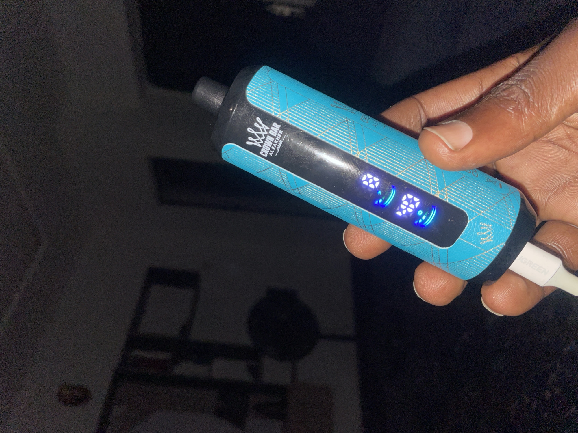An image captured in Abuja, Nigeria, features a close-up view of a hand holding a teal-colored vape device. The vape device has a black top and a digital display on its side that shows glowing blue numbers, indicating "78%". The device is adorned with a blue plaid pattern and features the "Crown Bar" logo and "Al Fakher" branding. A white charging cable is connected to the bottom of the vape. The background is dark and out of focus, with faint shadows of furniture and a circular object that may be a fan, suggesting an indoor setting. The lighting is dim, with the primary light source appearing to be the illuminated display on the vape device.