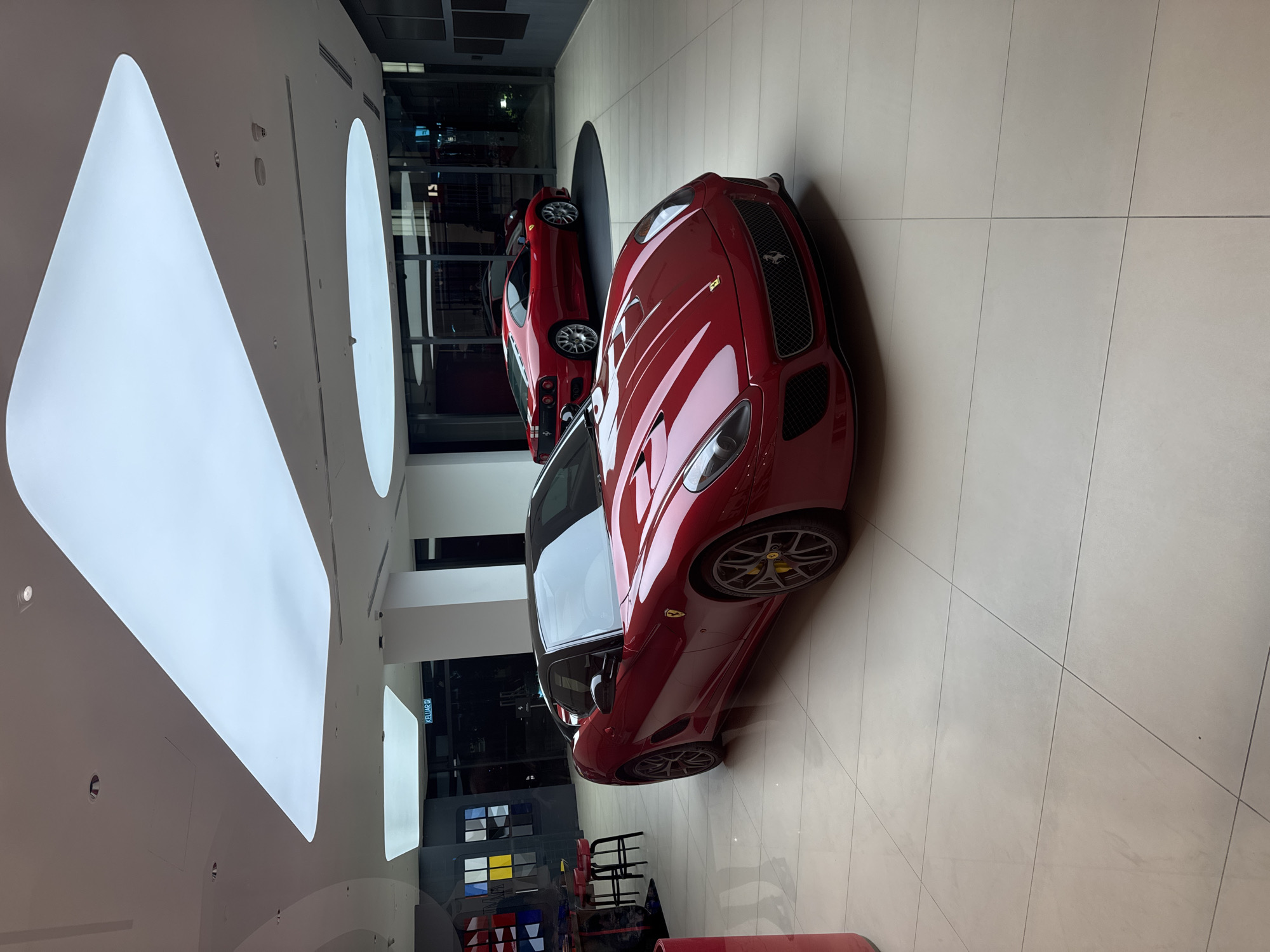 The image shows the interior of a retail space featuring two red Ferrari cars. The flooring is light-colored tiles with geometric patterns. The building appears modern with large, curved light fixtures on the ceiling and floor-to-ceiling glass walls. The location appears to be a car dealership or showroom in Kuala Lumpur, Malaysia. Additional furnishings include modern, colorful decor and seating.