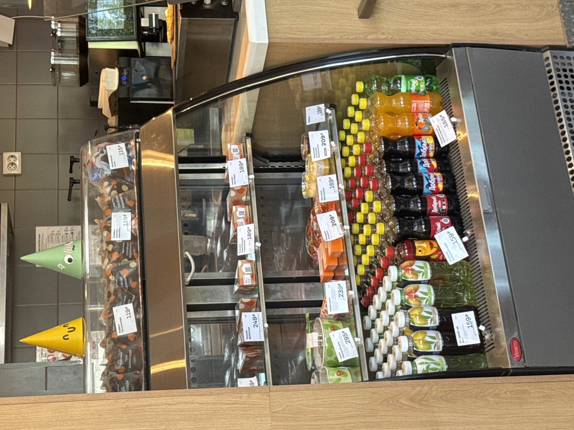 This image shows a refrigerated display case filled with various food and drink items, likely in a convenience store or small grocery shop. The display case is made of glass and metal, with shelves organized for optimal product presentation.
On the left side of the display, there are several packages of candies or chocolates, some with prominent labels indicating prices of "119p" and "249p". Two decorative, cone-shaped objects, one green with a cartoonish face and the other yellow with a similar design, are also visible in this section.
The main section of the display case is filled with bottles of beverages. There are rows of brightly colored drinks, including orange, yellow, and green liquids, as well as bottles of cola labeled with "Добрый" (Dobry). Prices such as "159p", "115p", "169p", "189p", "199p", "209p", "239p", and "295p" are visible on price tags. Some bottles have labels featuring fruit designs, like peaches and lemons, and some are labeled as "tea".
The setting appears to be indoors, with tiled walls visible in the background and a wooden countertop. The lighting suggests it could be daytime, but no specific time of day is discernible. The overall scene is one of a retail environment stocked with products ready for purchase. The presence of Russian text on the price tags and product labels ("Добрый", "Карамель", "Шоколада") indicates a Russian-speaking location.