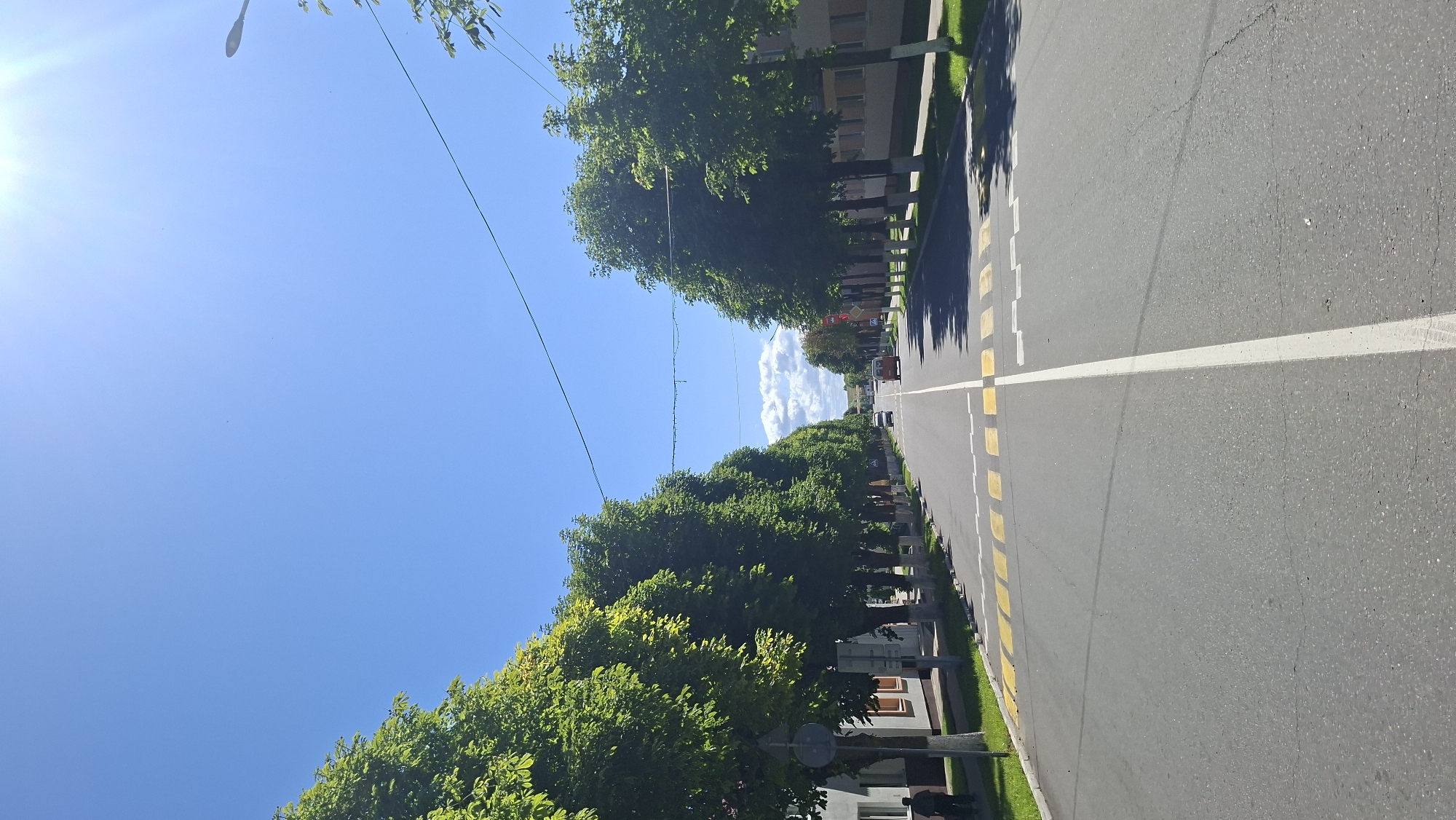 A paved road with yellow pedestrian crossing lines runs through a tree-lined street in Myadzyel, Belarus. The sky is clear and blue with the sun shining brightly. Trees with full green foliage line both sides of the road, casting shadows on the pavement. Buildings with beige facades and brown window frames are visible on the right side of the street. A few cars are driving on the road, including a white car and an orange van. A person is walking on the sidewalk on the left side of the street. The overall scene depicts a quiet, sunny day in a suburban or town setting.
