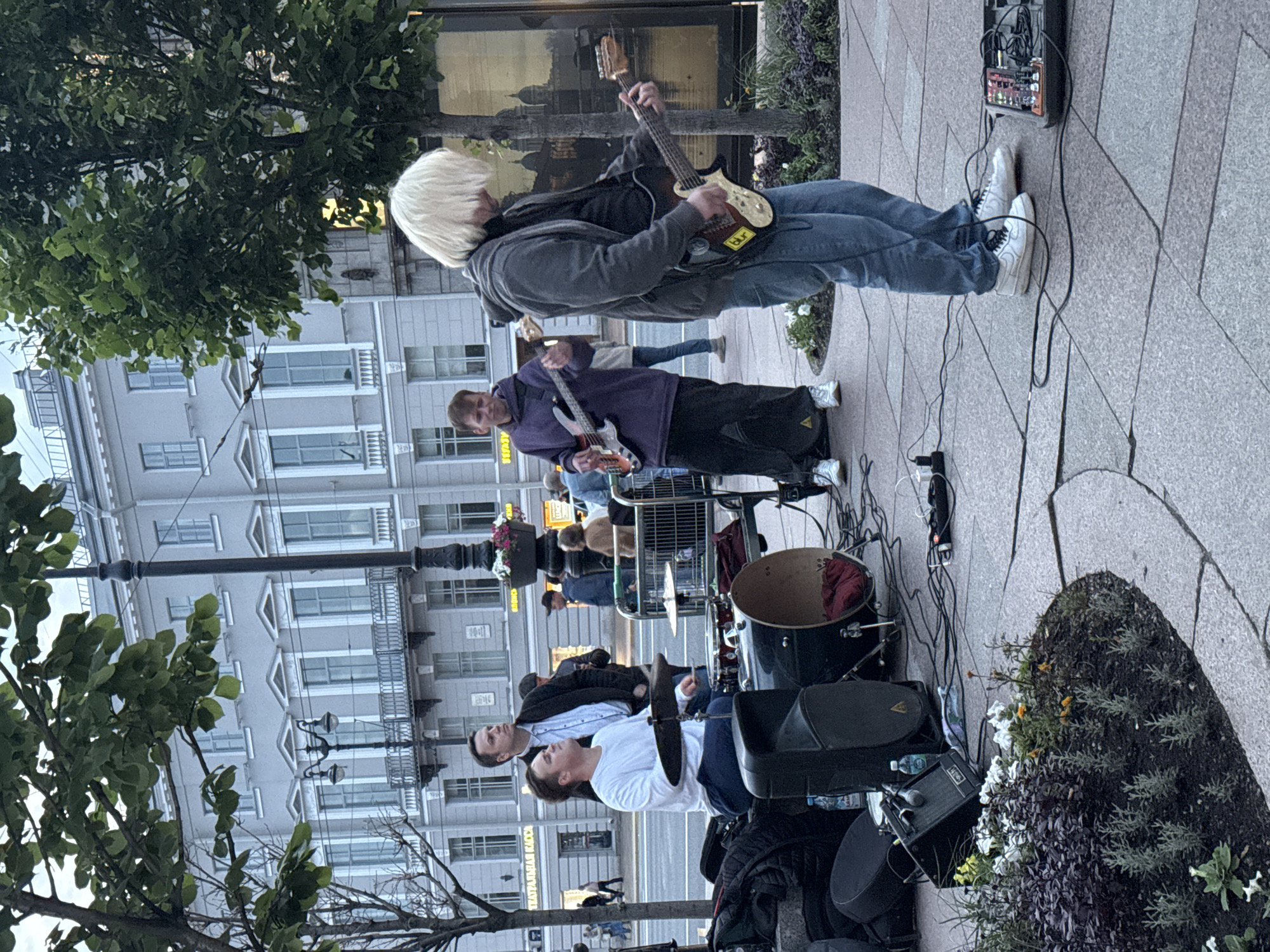 The image depicts a street performance by a band in Saint Petersburg, Russia, likely on Nevsky Prospect, given the architectural style of the buildings. Four individuals are visible, appearing to be part of a band. One person, with bleached blonde hair, is playing an electric guitar, wearing a grey hoodie and blue jeans. To their left, another person in a purple hoodie and black pants is also playing an electric guitar. A third individual, wearing a white t-shirt and blue pants, is seated at a drum kit. A fourth person, dressed in a dark jacket and light-colored shirt, stands behind the drummer, observing.
The band is set up on a paved area with a circular flower bed in the foreground. Musical equipment, including amplifiers, speakers, microphones, cables, and a pedalboard, is scattered around the musicians. The background features a large, ornate building with multiple windows and balconies, typical of St. Petersburg's classical architecture. Trees line the street, and a lamppost is visible. Other pedestrians can be seen in the distance, suggesting a public thoroughfare. The lighting suggests it is either early morning or late afternoon.