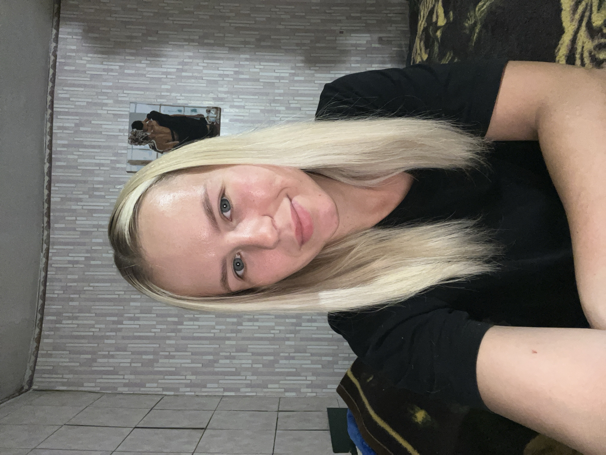 In an indoor setting, a young Caucasian woman with blonde hair is seen in a close-up, half-body shot, looking towards the camera. She is wearing a black t-shirt. The background consists of patterned wallpaper and a framed artwork depicting a couple. To the left and right of the woman, parts of a bed with a patterned blanket are visible. The lighting suggests an indoor environment.