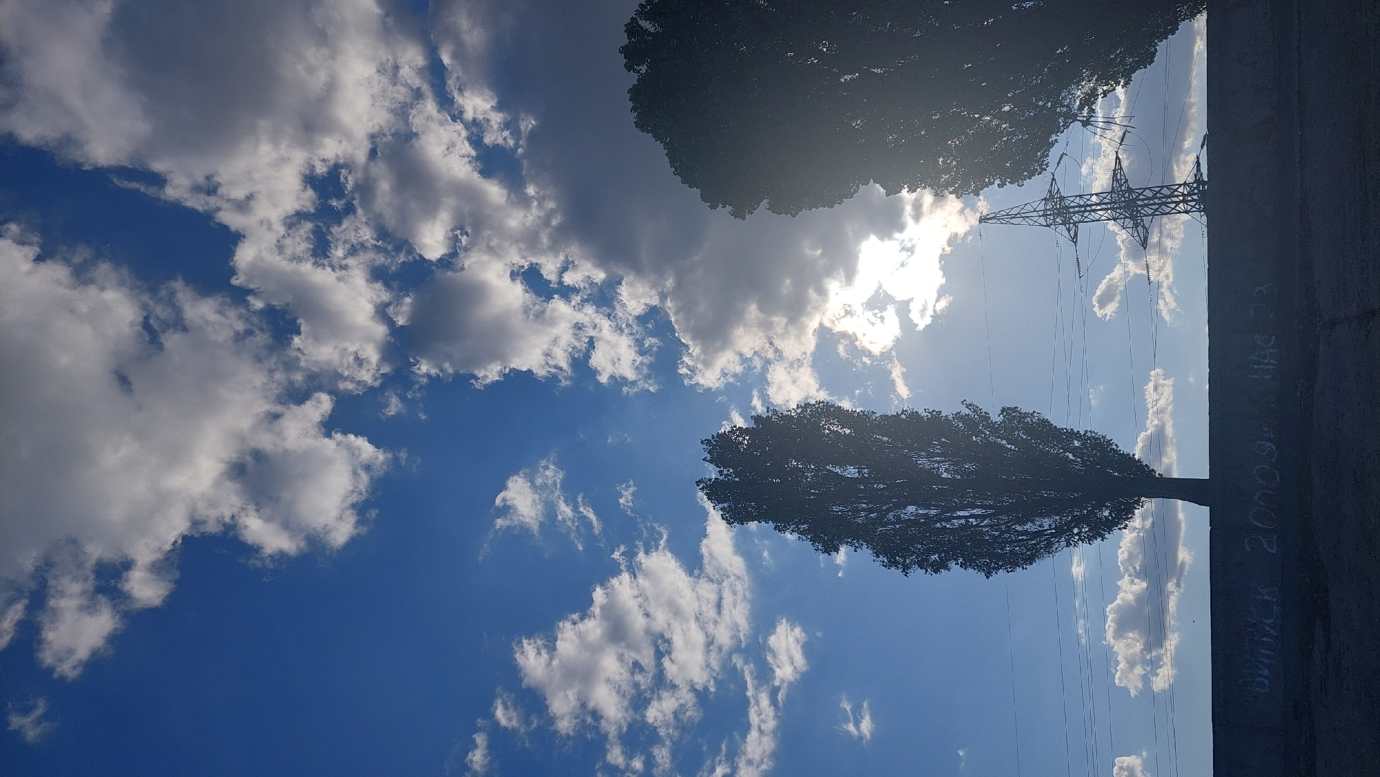 This image depicts a view of the sky with several trees and a power line tower in the foreground. The sky is a vibrant blue with a mix of white and grey clouds, suggesting either a partly cloudy or transitioning weather day. The sun is visible, partially obscured by clouds, casting a bright light that creates lens flare in the image.
Silhouetted against the sky are two tall, slender trees, one on the left and another on the right, framing the central portion of the scene. Between the trees, a power line tower with intricate latticework stands prominently. Several power lines are strung horizontally across the image, connecting to the tower and extending out of frame.
In the immediate foreground, a dark, flat surface serves as a base or wall, with faint white lettering or graffiti visible on its surface. The overall impression is an outdoor scene, likely in an urban or semi-urban environment, with infrastructure like power lines and a wall suggesting human presence. The context provided indicates this scene is located in Kozarovychi, Ukraine, a rural settlement in Kyiv Oblast.