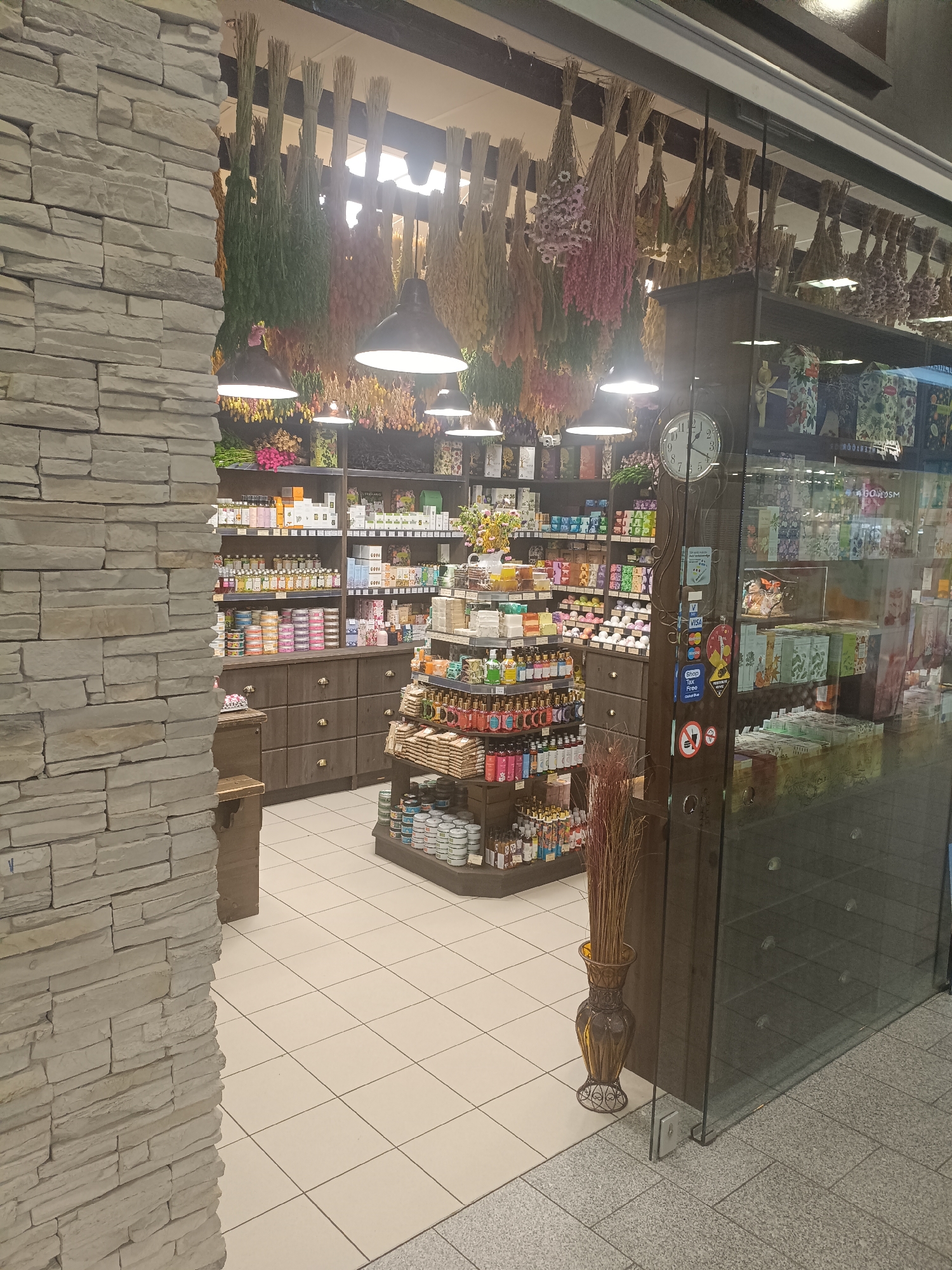 The image shows the interior of a shop that appears to sell natural products or gifts, located in Narva, Estonia. The shop's facade is made of stacked stone, and inside, shelves are filled with various items, including bottles, jars, and boxes. Dried herbs and flowers hang from the ceiling, creating a rustic ambiance. A large clock is visible on a display unit, and a decorative vase with dried branches stands near the entrance. The flooring is tiled. There are no people visible in the image. The scene appears to be a retail environment, likely a specialty store.