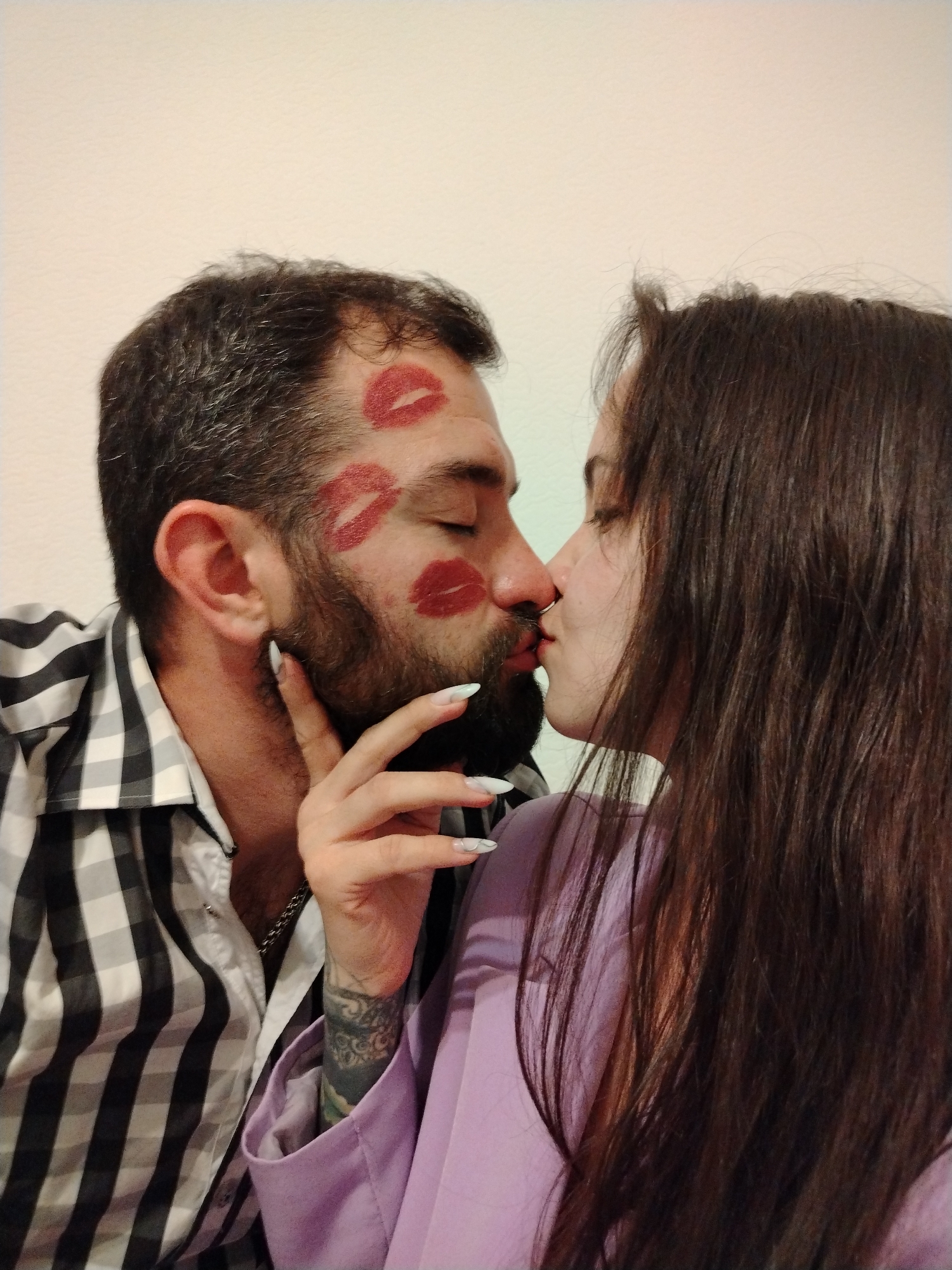 A close-up shot features a couple in an intimate moment. The man, with a salt-and-pepper beard, has several lipstick kisses on his face. He is wearing a black and white checkered shirt with a silver chain visible around his neck. The woman, with long dark hair, has one hand on the man's face, revealing a tattoo on her wrist and manicured nails. She is wearing a light purple top. The background is a plain white wall. The location is not specified as Noviy, Russia in the image. The interaction is a kiss.