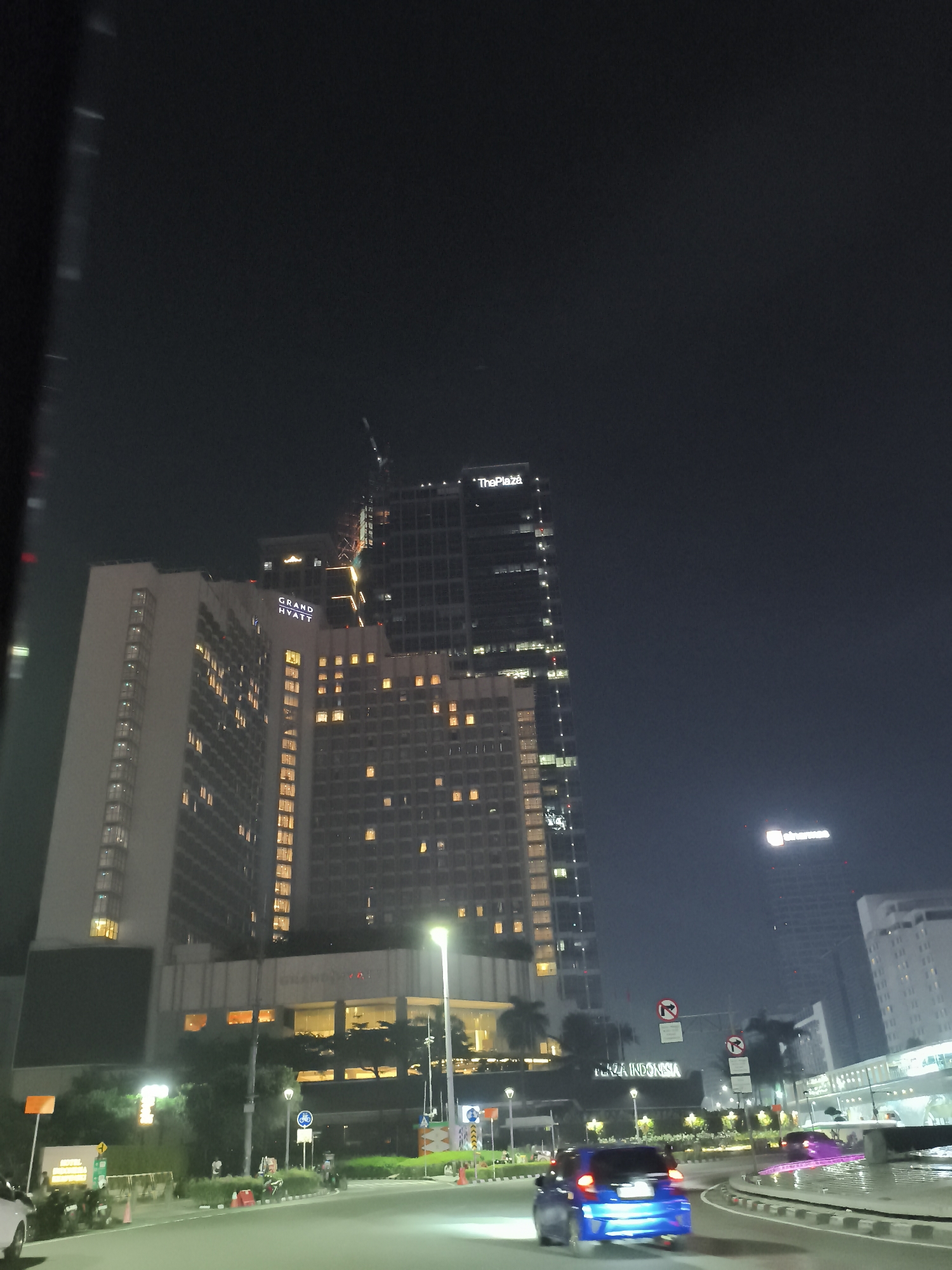 At night, the image shows Grand Hyatt and The Plaza buildings. A crane is visible atop one of the buildings. Traffic is flowing on the roads. There is signage for "Plaza Indonesia" in the background.
