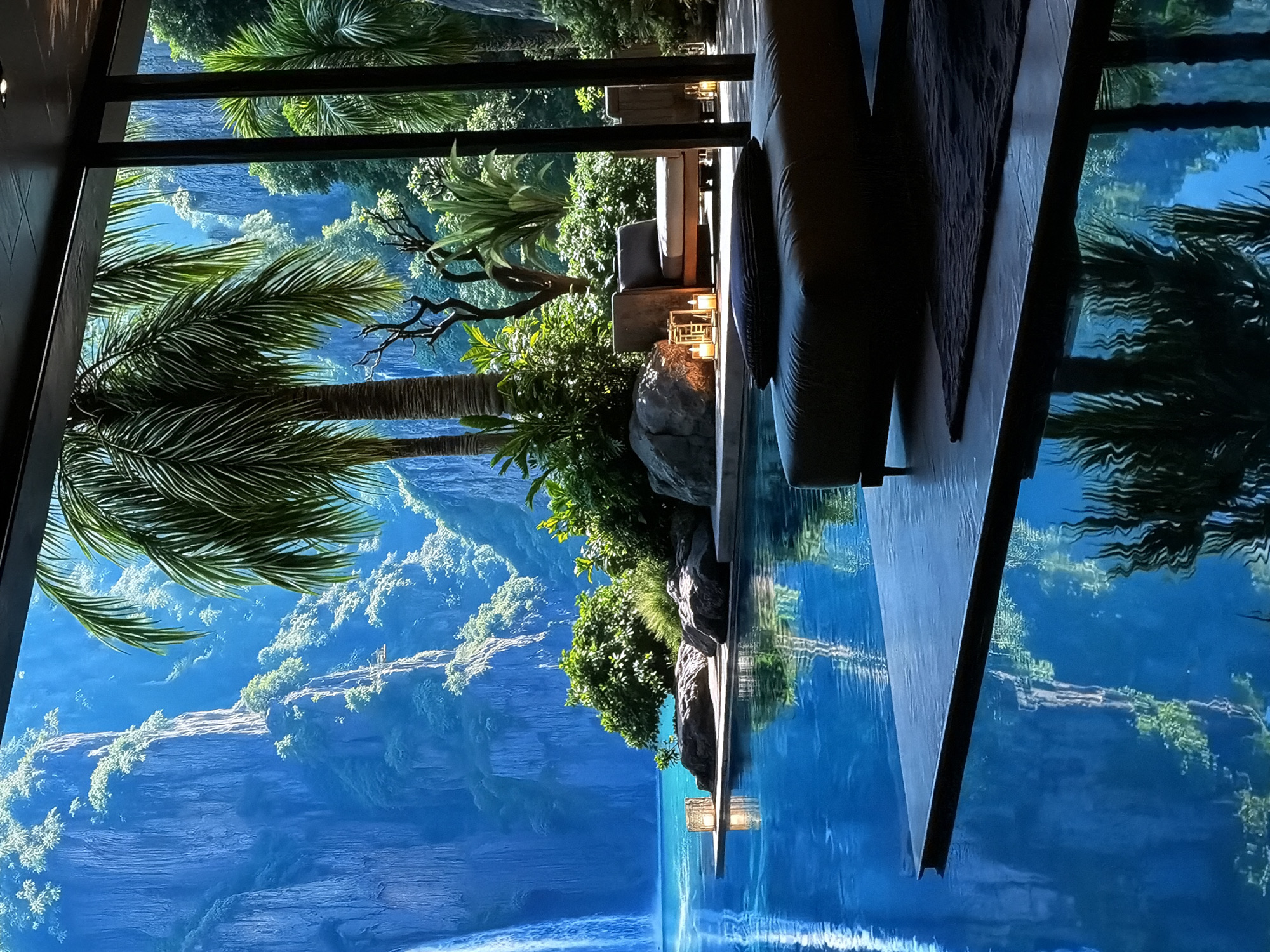 The image captures a luxurious and serene setting, likely a high-end lounge or resort area, with a dramatic panoramic view. In the foreground, to the right, a comfortable outdoor seating arrangement is visible, featuring dark-colored sofas and cushions. Decorative lanterns or candle holders with lit candles are placed on a low table, suggesting a cozy evening ambiance.
To the left, lush tropical foliage, including prominent palm fronds, frames the expansive vista. The view beyond appears to be a vast, vibrant landscape featuring towering, rugged blue mountains or cliffs covered with patches of bright green vegetation. Directly adjacent to the seating area, a large, calm body of water (possibly an infinity pool or a reflective surface) perfectly mirrors the intense blue sky and the distant mountain range, creating a seamless and immersive illusion. The reflections are crisp and deep, enhancing the sense of tranquility.
The overall scene has a highly stylized, almost hyperrealistic quality, particularly the distant landscape with its vivid blues and greens. This visual characteristic, combined with the stated location context of Sofiivska Borshchahivka, Ukraine (which is not known for tropical mountain landscapes), suggests that the distant view might be a very large high-resolution digital display, a projection, or a digitally rendered backdrop rather than a natural outdoor environment. No people are visible, and there is no visible text. The time of day appears to be late afternoon or early evening, as indicated by the lit candles, though the distant view is brightly illuminated.