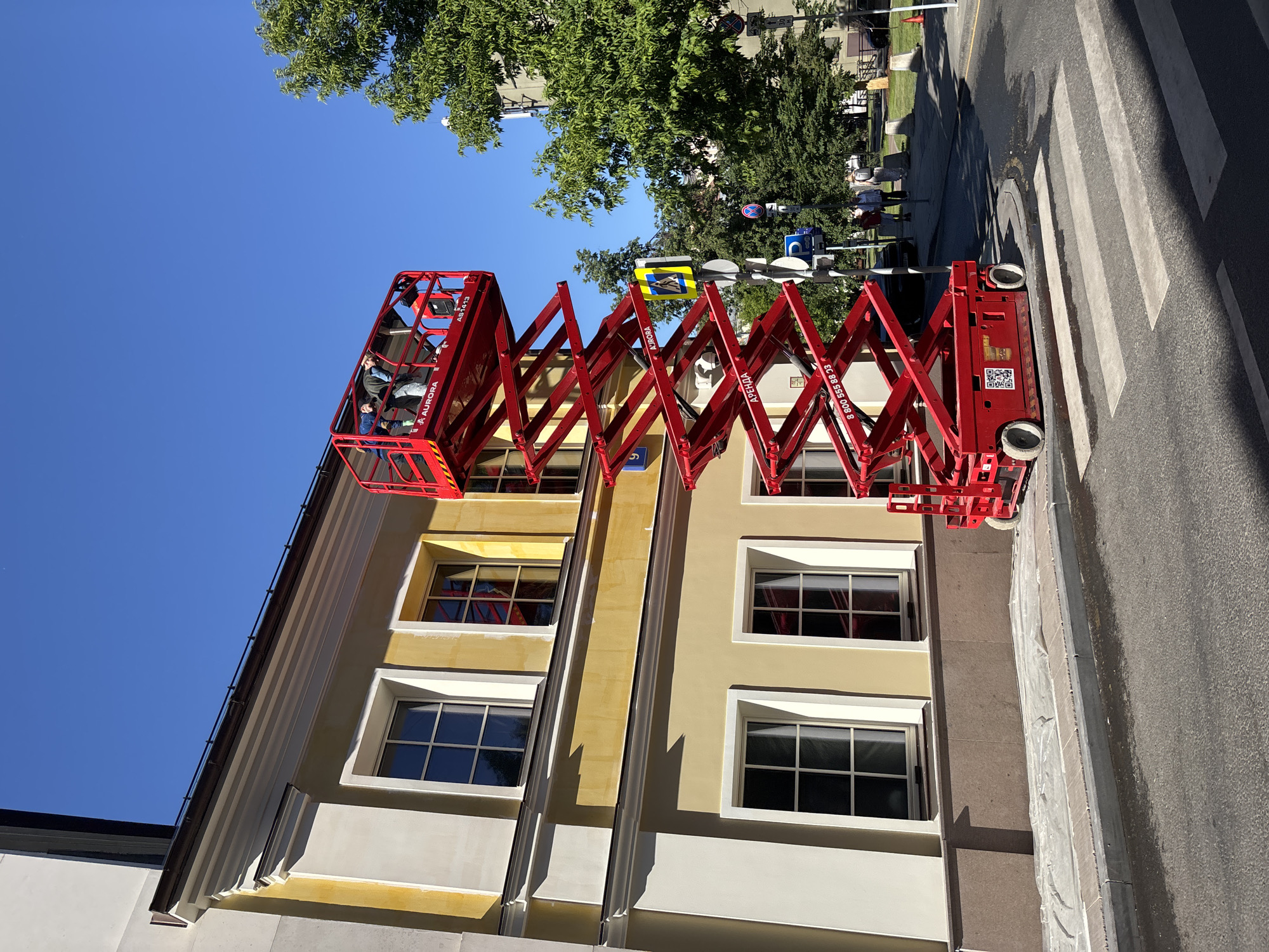 A red Aurora AS1413 scissor lift is extended to its maximum height against the facade of a multi-story building on a clear, sunny day in Moscow, Russia. Four individuals are positioned within the lift's safety cage at the top, appearing to engage in work on the upper portion of the building.
The building features a pale yellow facade with white classical architectural details, including window frames, cornices, and pilaster-like elements. Multiple windows with white frames and dark panes are visible across two stories. The lowest visible section of the building has a darker, possibly granite or stone, base. At ground level, the sidewalk directly in front of the building is covered with clear plastic sheeting, suggesting ongoing construction or renovation work. A dark blue street number plate displaying "9" in white is affixed to the building's left side.
The scissor lift itself is bright red, with "AURORA" branding and the model number "AS1413" visible on the platform. The word "АРЕНДА" (RENT) and a Russian toll-free phone number "8 800 555 88 33" are printed on the lower, stationary section of the lift. A QR code is also present on the base.
The scene includes an asphalt road in the foreground with a clearly marked white pedestrian crosswalk. A concrete curb separates the road from a paved sidewalk. Various street signs are mounted on poles to the right of the lift: a yellow diamond-shaped sign with a black pedestrian symbol, a blue circular sign with a white 'P' indicating parking, and another sign further down, partially obscured, showing a number "30" which may denote a speed limit or other restriction. Lush green trees are visible along the street and in the background under a vibrant blue sky. Several pedestrians are visible on the sidewalk further down the street.