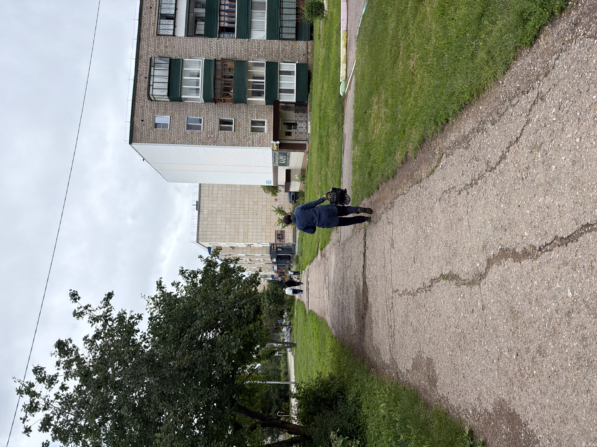 A person in a dark blue jacket walks away from the camera on a cracked concrete sidewalk. The sidewalk is bordered by a strip of green grass, and beyond that are multi-story buildings. The buildings appear to be residential apartment blocks, with several windows visible. One building is lighter colored and features a white, corrugated siding. Another is constructed of brick and has green trim around some of its windows. Trees and greenery are also present in the scene. The sky is overcast and cloudy. The time of day appears to be daytime. There is some visible text on a sign attached to one of the buildings, reading "ПЕРМСКИЙ КАРЕ" and another sign below it which is partially obscured.