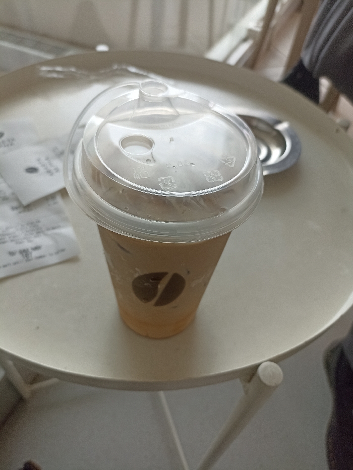 A disposable cup of iced coffee with a plastic lid sits on a white round table with a white metal leg. The coffee cup has a brown logo with a coffee bean shape on its side. On the table, there are several crumpled pieces of paper and a partially visible silver tray, likely from a coffee shop, indicating that the image was taken indoors. The setting is consistent with a café in Purwokerto, Indonesia, based on the provided coordinates (-7.42, 109.245). The image is a simple, unassuming snapshot of a cup of coffee on a table.
