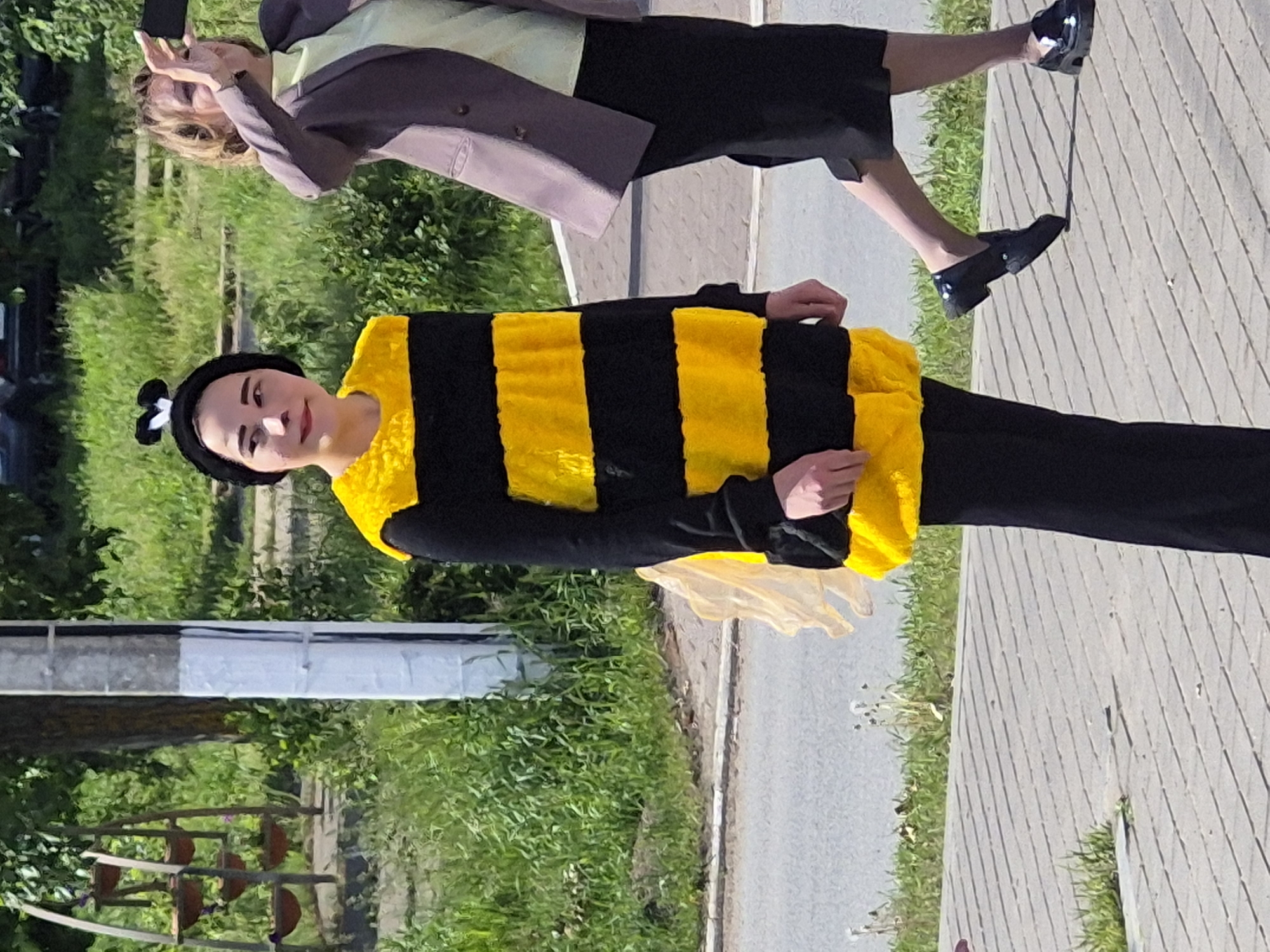 A woman dressed in a bee costume stands on a paved path, next to a grassy area, in what appears to be a public space in Chaikovsky, Russia. The bee costume is yellow and black. Another person is partially visible in the upper portion of the image, taking a picture with a phone. The location includes benches.