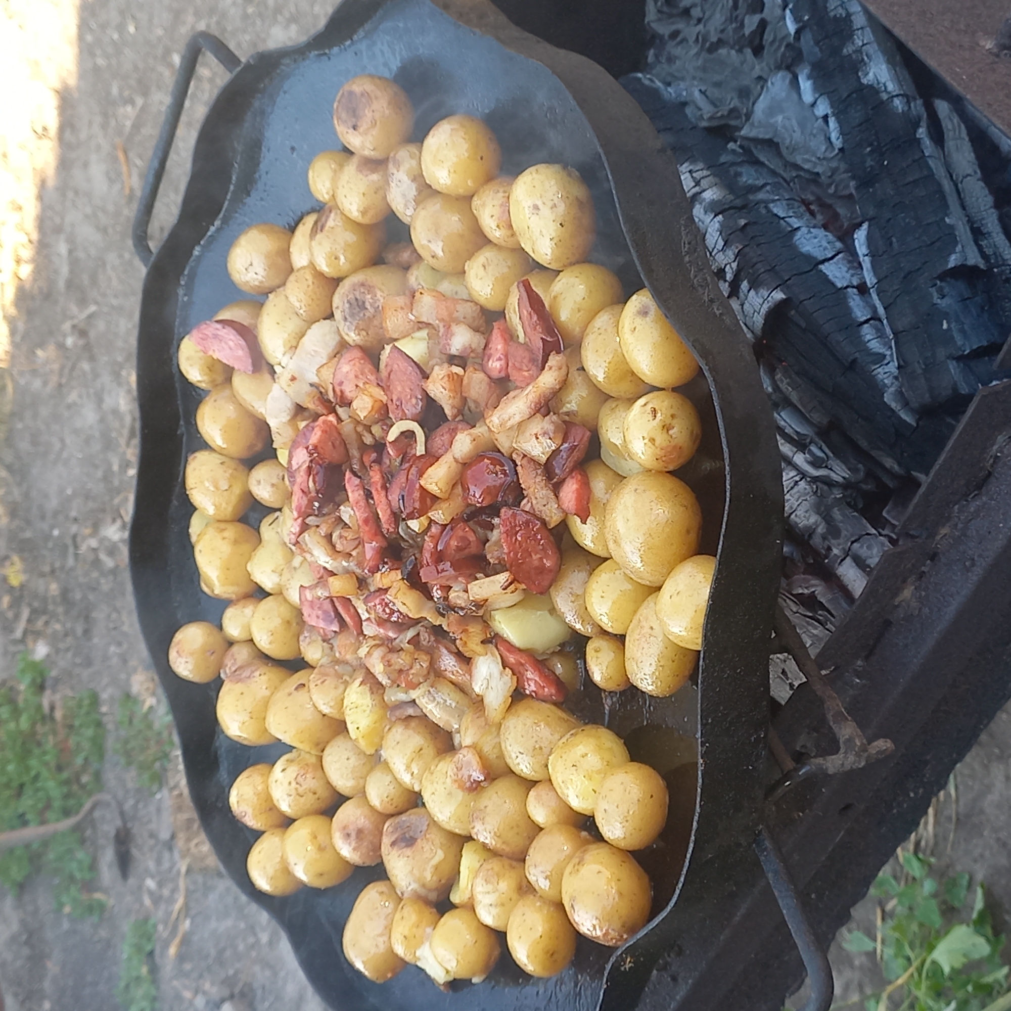 In Klyntsi, Ukraine, a large, black, round cast-iron pan is placed over a bed of glowing embers and charcoal. The pan is filled with small, whole, yellow potatoes, many of which appear slightly browned or charred from the heat. Mixed in with the potatoes are pieces of cooked sausage, some appearing sliced and others in smaller, irregular chunks. A scattering of what looks like fried onions or other vegetables can also be seen among the potatoes and sausage. The scene is outdoors, with a blurry background of dry earth and green foliage, suggesting a rustic or campsite setting. The pan has two dark handles, one of which is partially visible on the right side. The cooking is actively taking place, as indicated by the steam or smoke rising from the food.