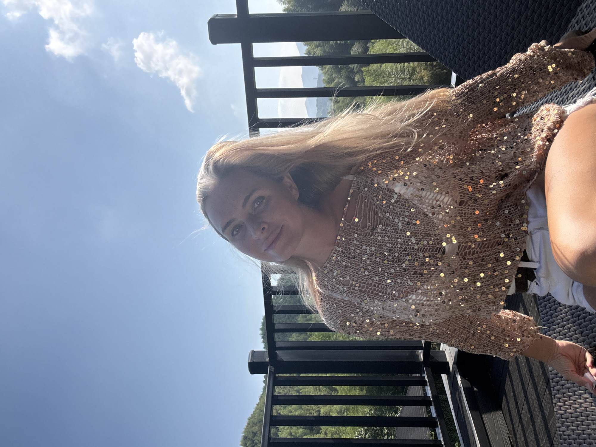 A woman with blonde hair is sitting on a chair outdoors on a balcony. She is wearing a sequined, open-knit sweater over a white bikini top and white shorts. She is looking at the camera with a slight smile. The background shows a railing, trees, and mountains. The sky is blue with some clouds. The image is taken on a sunny day.