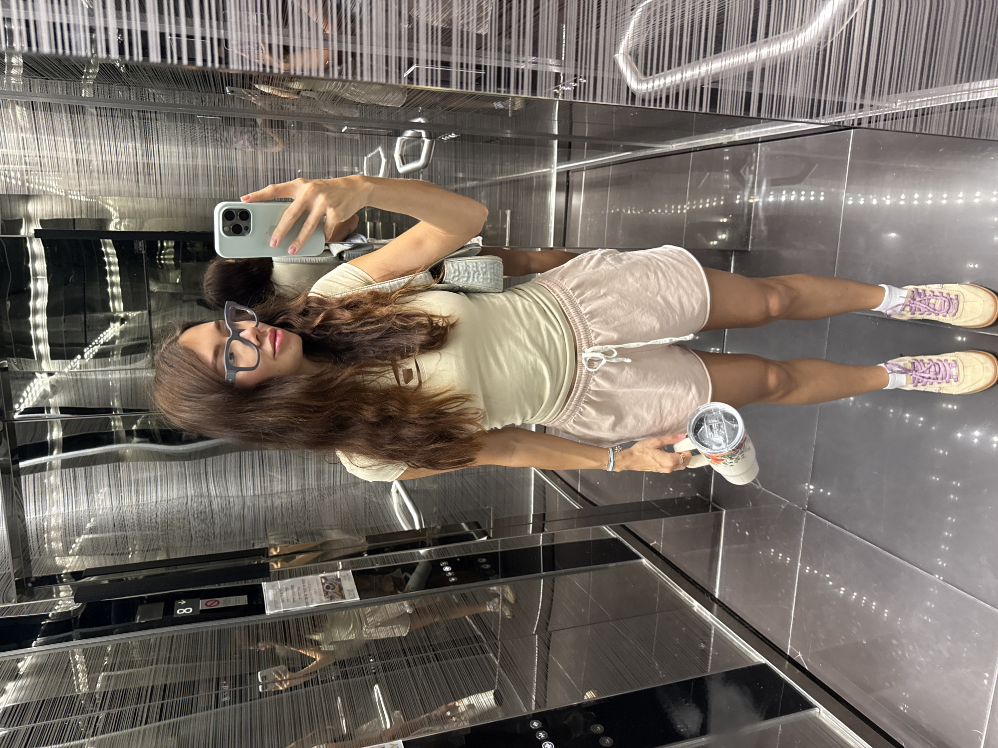 A woman is taking a selfie in an elevator. She is holding a cup in her left hand and a phone in her right hand. She is wearing glasses, a light yellow shirt, pink shorts, white socks, and tennis shoes. The elevator has mirrored walls and a panel with buttons. The image is taken in an elevator of a location in the city of Unknown, Unknown.