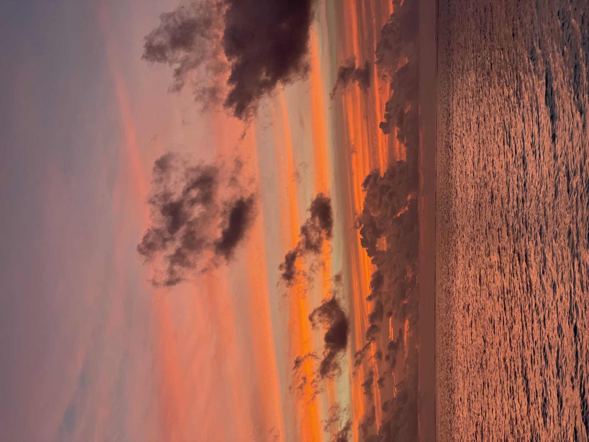The image depicts a sunset over a body of water, with the sky filled with vibrant orange, pink, and purple hues. Several dark clouds are scattered across the sky, contrasting with the bright colors. The water is calm and reflects the colors of the sky, creating a serene and picturesque scene. The location is not specified in the city of Unknown, Unknown.