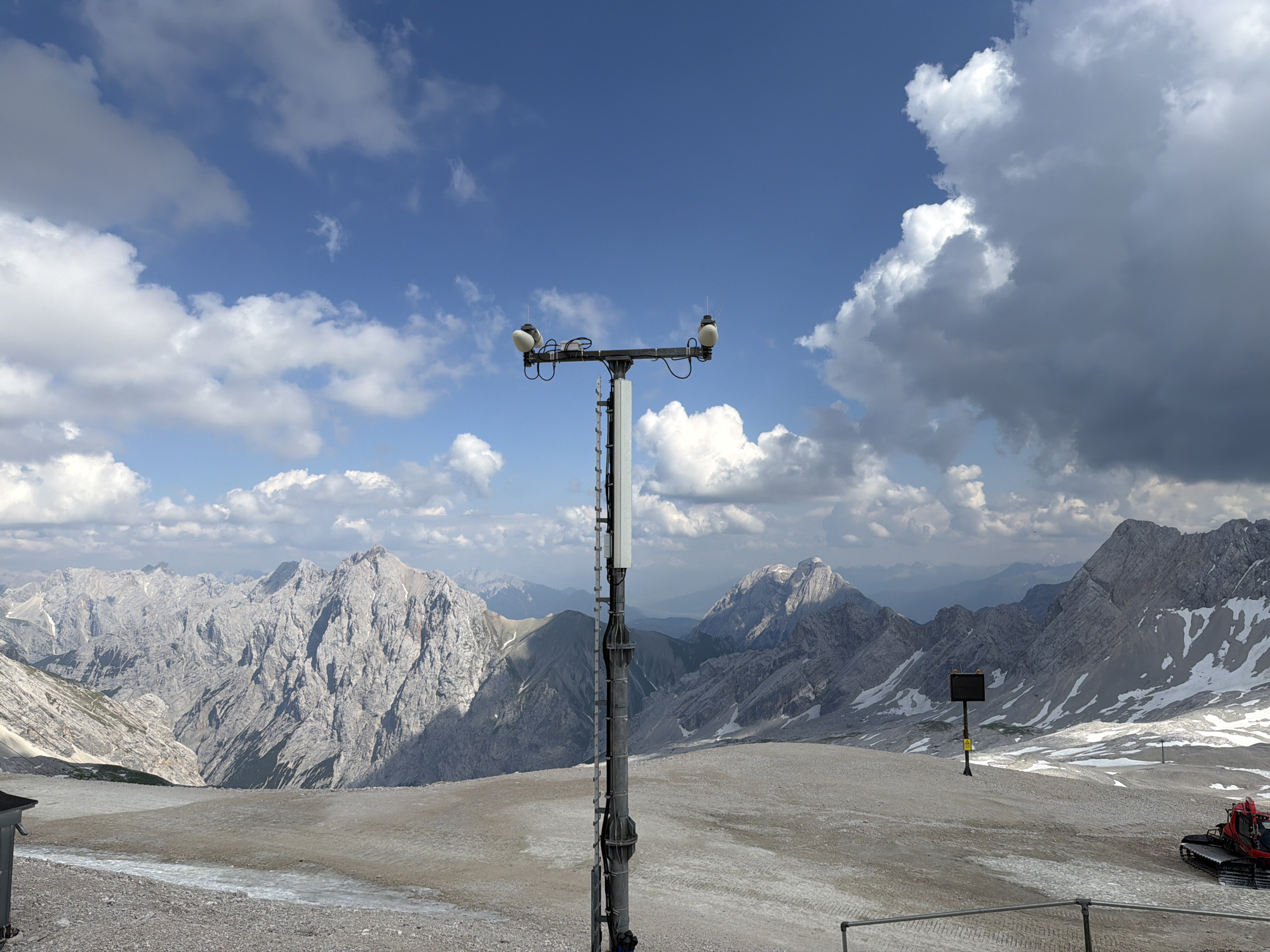 This media file captures a panoramic view of a mountainous landscape under a partly cloudy sky. The foreground features a weather station with two white spherical sensors mounted on a tall metal pole. The station is situated on a gravelly, uneven terrain. In the background, rugged, rocky mountains dominate the scene, with patches of snow visible on their slopes, suggesting a high-altitude location. The sky is a vibrant blue, interspersed with fluffy white and darker gray clouds, hinting at a dynamic weather system.
To the right of the frame, a red snow groomer is parked on the barren ground, indicating the possibility of snow sports or maintenance activities in the area. A dark, square signpost with a yellow warning symbol is also visible in the mid-ground. The overall impression is one of a vast, natural environment, likely a mountain peak or high plateau, accessible to visitors or workers. The lighting suggests it is daytime, possibly late morning or early afternoon, given the brightness and the shadows cast by the mountains.
The location appears to be Garmisch-Partenkirchen, Germany, a region known for its alpine scenery and outdoor activities. No people are visible in the frame, focusing the attention on the landscape and the technological equipment. The atmosphere is one of natural grandeur and technological presence, coexisting in a remote, elevated setting.