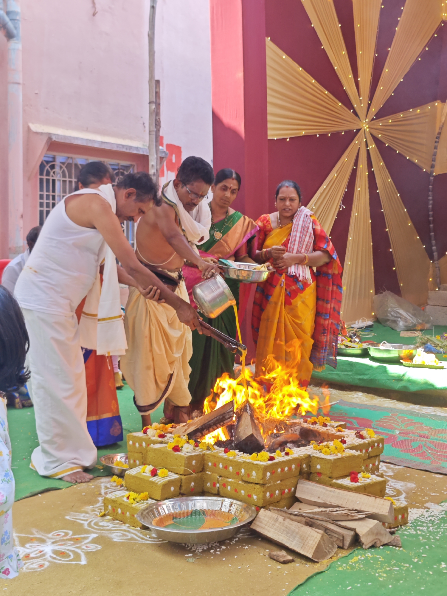 A religious ceremony is underway outdoors, with a group of people gathered around a small, stacked arrangement of bricks forming a structure for a fire. The bricks are decorated with small yellow and red flowers. A fire burns brightly within the brick structure, with logs placed around it. Several people are involved in the ritual. One man, wearing a white dhoti and a white cloth draped over his shoulder, is pouring liquid from a metal container into the fire. Another man, dressed in a dhoti and a white sacred thread, stands beside him, also holding a metal container. Two women in colorful saris are observing the ceremony. One woman in a green sari stands behind the men, while another woman in a yellow and red checkered sari stands to the right, holding a metal tray. The background shows a building with pink and maroon walls and a decorative fabric display with lights. The ground is covered with a green cloth and a yellow mat, with rice grains scattered around. The scene appears to be taking place in Puttaparthi, India, suggested by the attire and the context of a Hindu ritual.