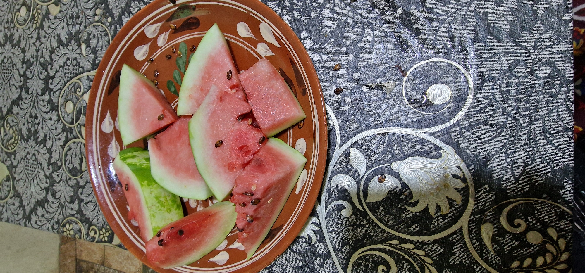 A close-up, overhead shot features a brown, rustic ceramic plate filled with sliced watermelon. The watermelon is cut into triangular wedges, some with rind still attached, displaying the vibrant pink flesh speckled with dark seeds. A few stray watermelon seeds are scattered on the patterned tablecloth beneath the plate. The tablecloth is dark gray with an intricate damask-like pattern in a lighter gray and silver, featuring floral and swirling motifs. The lighting is dim, casting soft shadows. The scene is likely indoors, given the tablecloth and what appears to be a tiled floor in the background. There is no indication of a specific location in Karshi City, Uzbekistan, or any people present.