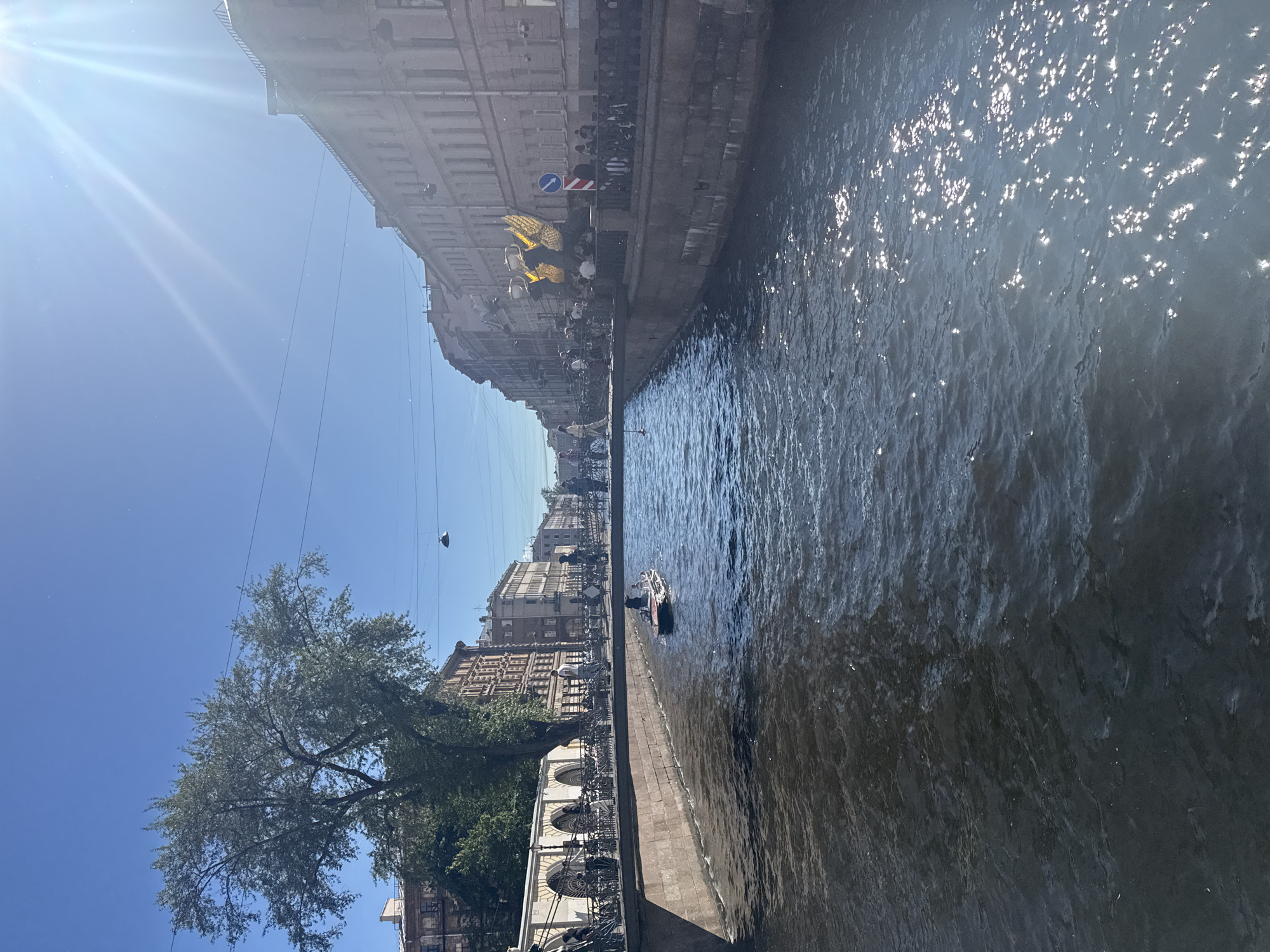 The image depicts a canal in Saint Petersburg, Russia, under a bright, sunny sky, suggesting midday. The water, shimmering with sunlight, reflects the overcast sky and the ornate, historic buildings lining the canal. A single, small boat with people aboard navigates the canal, creating ripples on the water's surface. The architecture of the buildings is detailed, with multiple windows and classical embellishments, hinting at the city's rich history. A lush green tree extends its branches towards the canal on the left side of the frame, adding a touch of nature to the urban landscape. Overhead, power lines crisscross the sky, a common sight in urban environments. The scene is peaceful, with no indication of any particular event. The overall impression is one of a pleasant day in a picturesque European city.