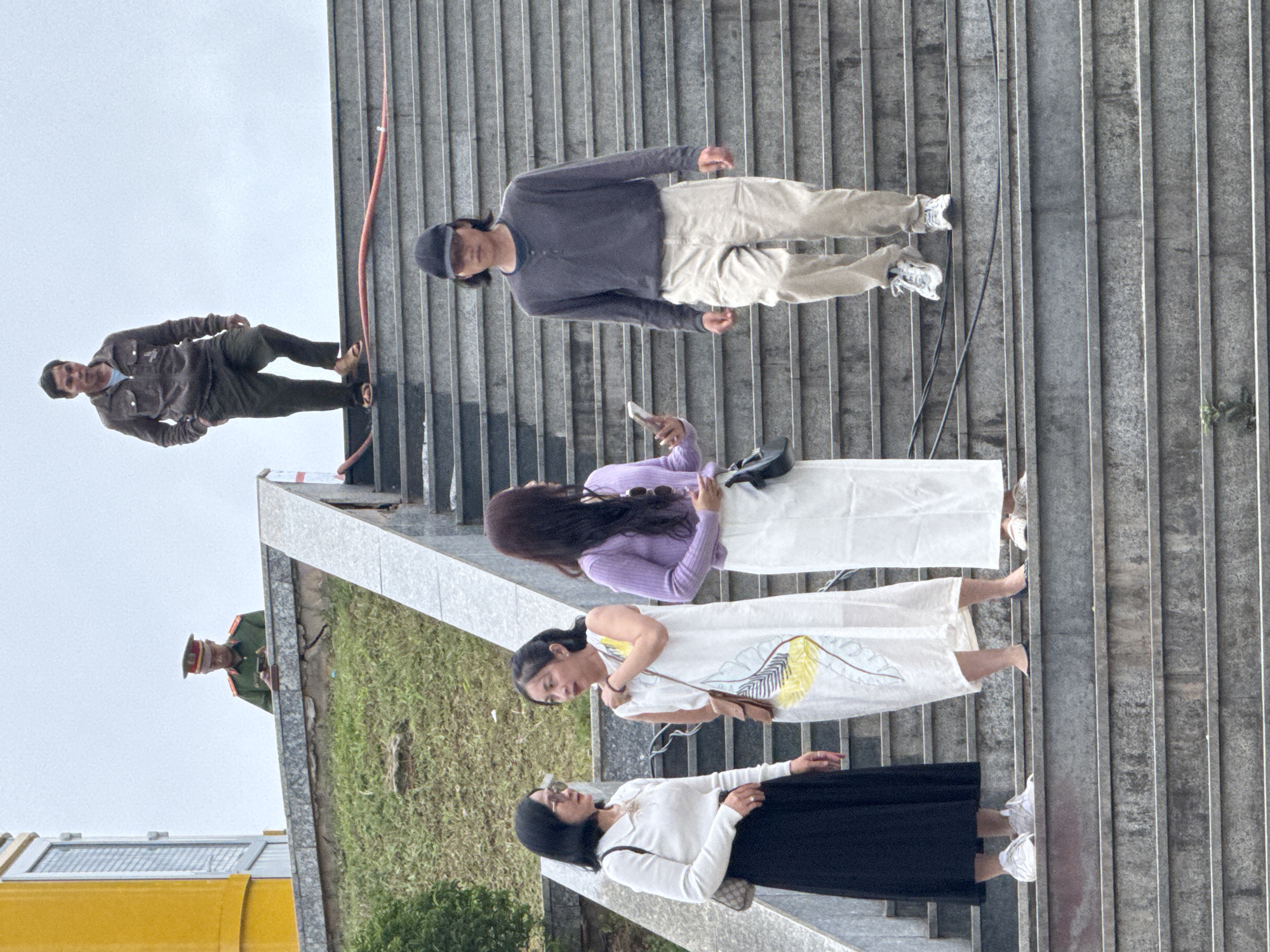 This media file captures a moment on a set of outdoor stairs, likely in Da Lat, Vietnam, given the context. Several people are present, with some ascending or descending the stairs.
In the foreground, two women are visible. One woman, wearing a white long-sleeved shirt and black skirt, is looking down. The second woman, in a white dress with a yellow and green floral pattern, is also looking down. Further up the stairs, a third woman, dressed in a purple sweater and white skirt, is holding a phone and looking towards her left.
Above them, a man in a dark long-sleeved shirt and light-colored pants is standing on the stairs, facing away from the camera. To his left, another man in a brown jacket and dark pants is walking down the stairs.
In the background, on a grassy embankment to the left, a person in a military-style uniform with a hat is partially visible, looking towards the right. A yellow structure, possibly a building or vehicle, is also present in the lower left corner.
The weather appears overcast, contributing to a diffused light. The setting is urban or semi-urban, with the prominent feature being the concrete stairs. The people seem to be engaged in everyday activities, with no specific event clearly indicated. The overall mood is casual. No visible text is present in the image.
