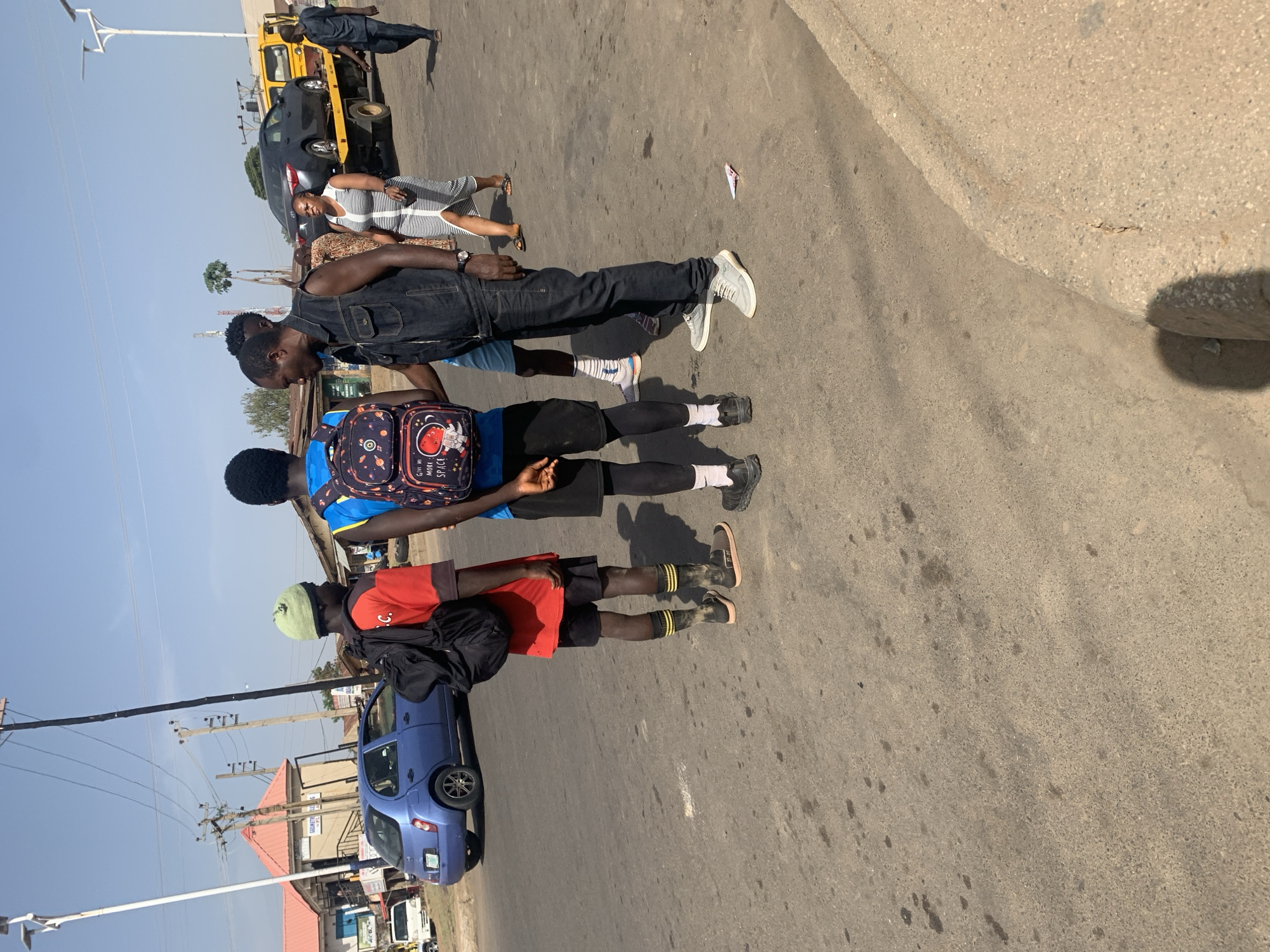 The image captures a street scene in Rafin Par, Nigeria. Three young men walk along the shoulder of a paved road. The first individual is dressed in a denim vest and black pants, wearing light-colored sneakers. The second wears a blue t-shirt with black shorts and carries a backpack with space-themed graphics. The third individual dons a red t-shirt, black shorts, and wears a light green cap, also carrying a backpack. A blue car is parked on the side of the road near the third individual. In the background, other vehicles including a tow truck, buildings, and utility poles are visible. The sky is clear.