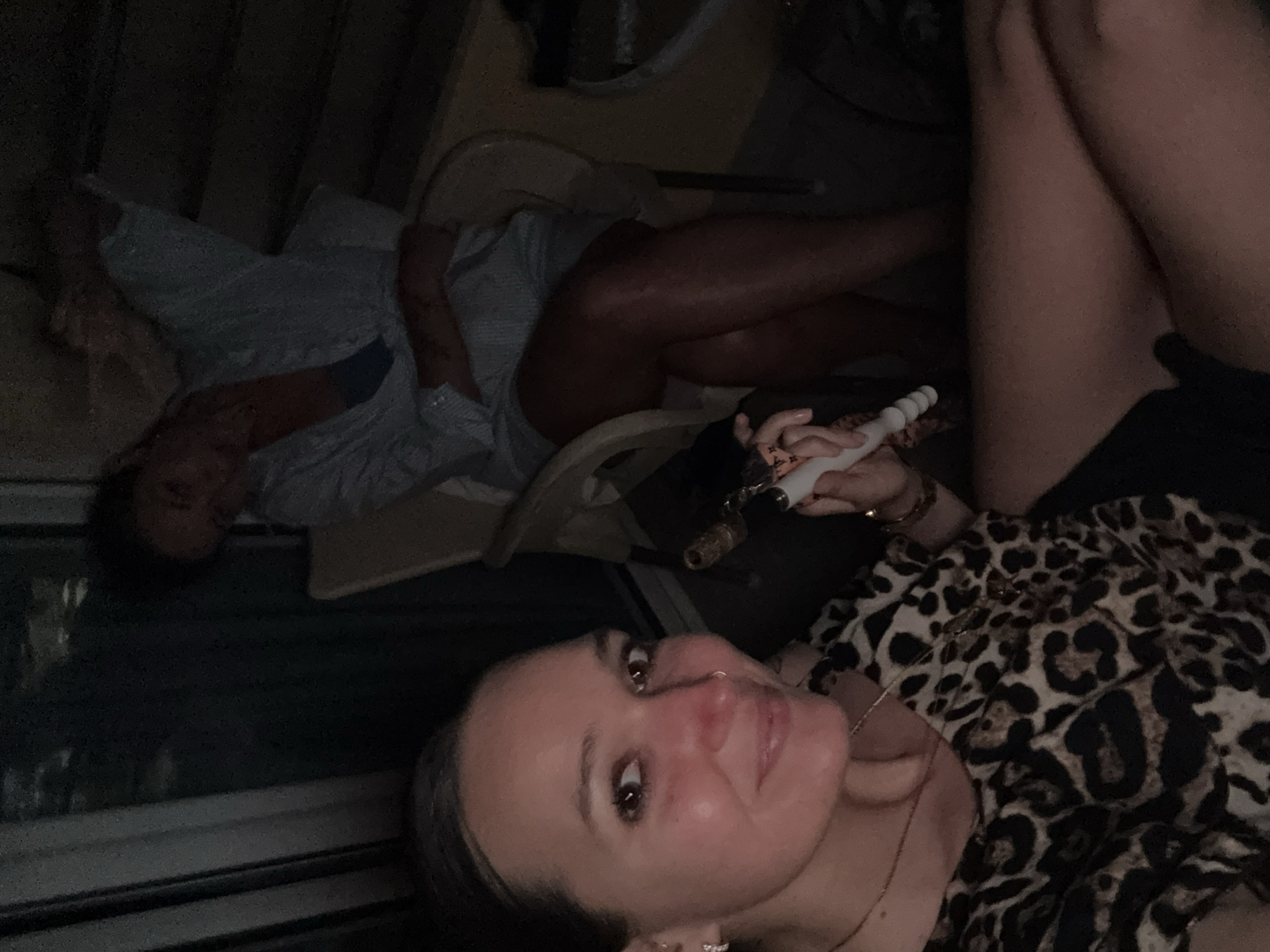 Two women are present in the image, seemingly on a balcony or outdoor seating area. The woman in the foreground, on the left, is facing the camera and smiling. She has dark hair pulled back, brown eyes, a nose ring, and is wearing a leopard-print top and gold jewelry. She is holding a white cylindrical object with a textured grip, possibly a vape pen or similar device. The woman in the background, on the right, is seated in a chair and appears to be looking away from the camera. She is wearing a light blue button-down shirt and has her legs crossed. The environment is dimly lit, suggesting it is either evening or night. The background shows some structural elements and foliage, indicating an outdoor setting, possibly within a residential or hospitality complex. The overall impression is a casual, relaxed social gathering. The location is identified as Budva, Montenegro.
