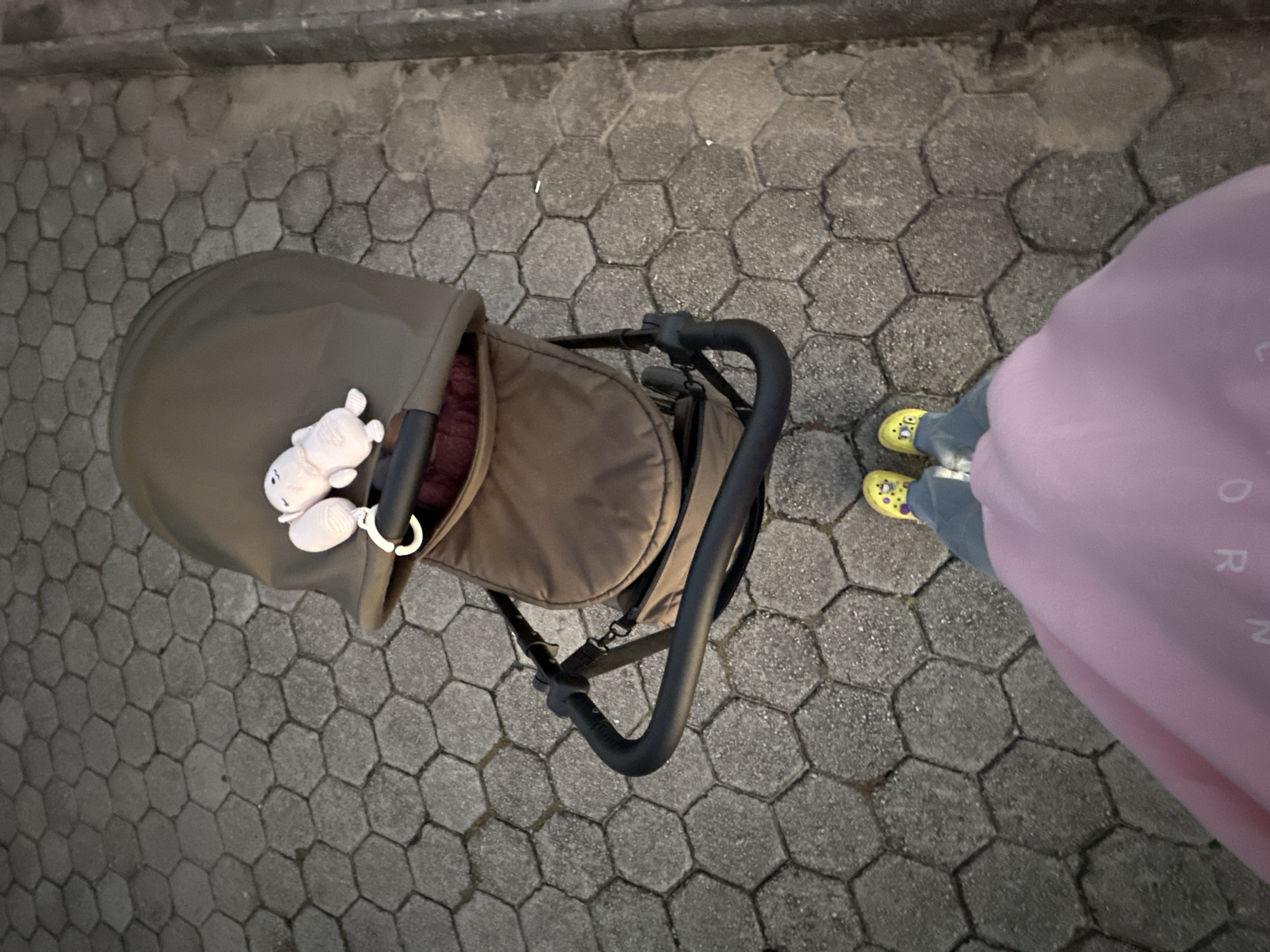 The image shows an overhead shot of a baby stroller on a gray hexagonal paved surface. The stroller is primarily brown with a black handle and a white toy attached. To the right of the stroller, a person is wearing yellow Crocs and a pink outfit with "UNICORN" lettering. The location appears to be an urban area in Florianópolis, Brazil, judging from the pavement style commonly used in the city.
