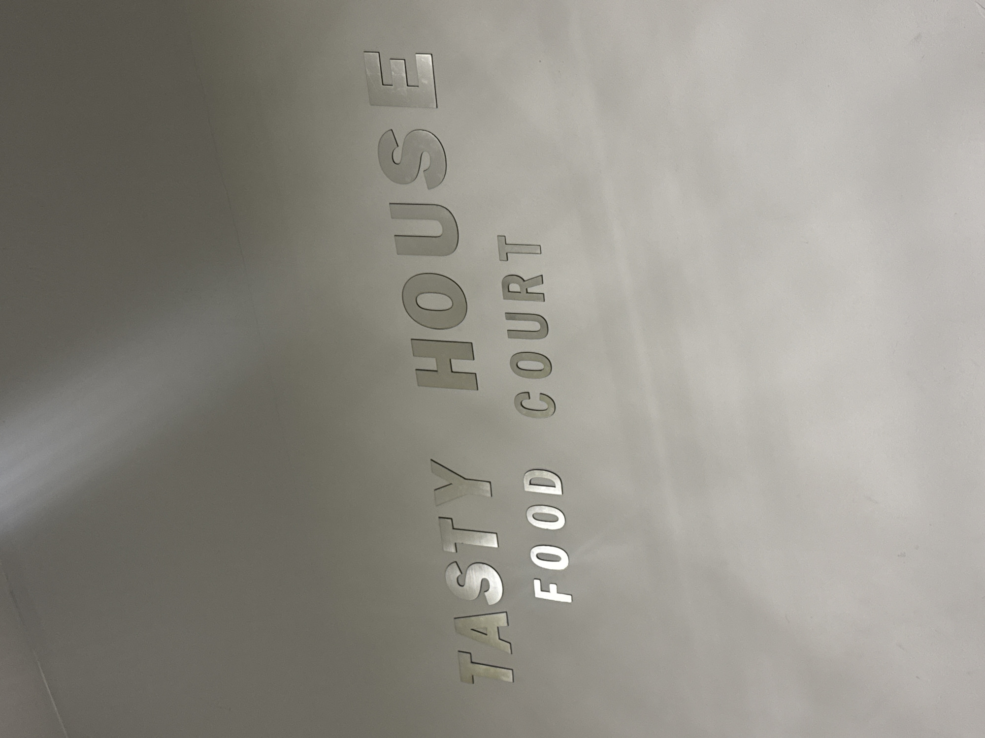 The image displays a close-up of a sign for a food establishment, likely located in an indoor commercial setting such as a shopping mall or building in Yerevan, Armenia. The sign features raised, metallic silver letters against a plain, light grey background.
The text on the sign is oriented vertically and reads "TASTY HOUSE" on the upper line, and "FOOD COURT" on the lower line. The letters appear to be embossed or made of a reflective material, catching the light and creating a subtle three-dimensional effect. The background is a smooth, uniform surface, possibly a painted wall or ceiling, with slight variations in shading indicating ambient indoor lighting.
No people or activities are visible in the frame, focusing solely on the sign itself. The design of the sign is clean and modern.