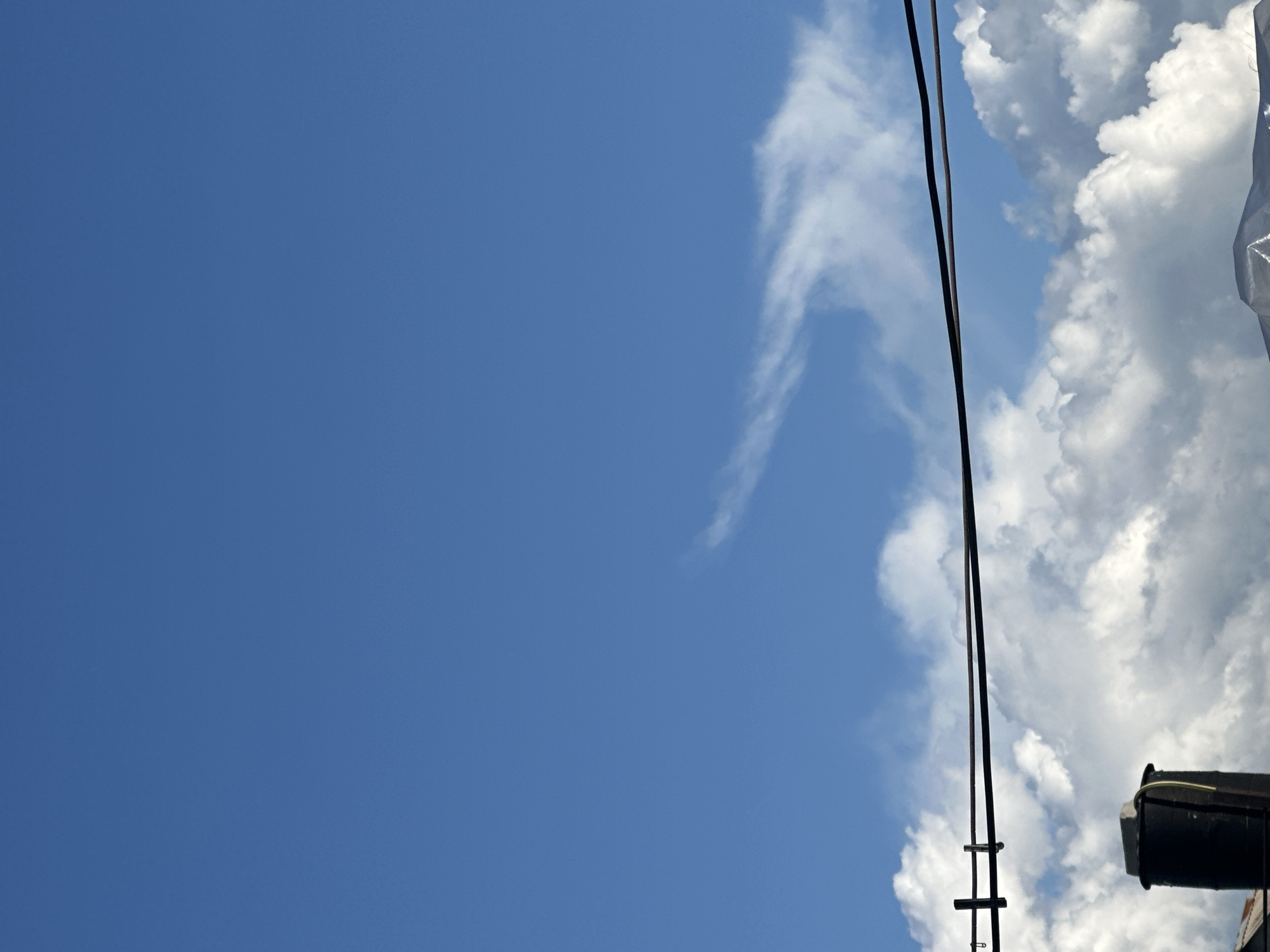 The media file is a photograph taken from a low angle, looking upwards towards a bright blue sky. The sky is mostly clear, with a few wispy white clouds scattered across it. On the right side of the frame, there are large, puffy white clouds, suggesting fair weather. Two thin, dark wires run vertically through the center of the image, intersecting with a ladder-like structure. To the far right, a dark, cylindrical object, possibly a part of a building or industrial equipment, is partially visible. The lighting indicates it is daytime, with the sun high in the sky. There is no discernible activity or event taking place, and no text is visible. The overall impression is one of a clear, open sky with some man-made elements intruding into the natural scene. The location is specified as Tobyl, Kazakhstan.
