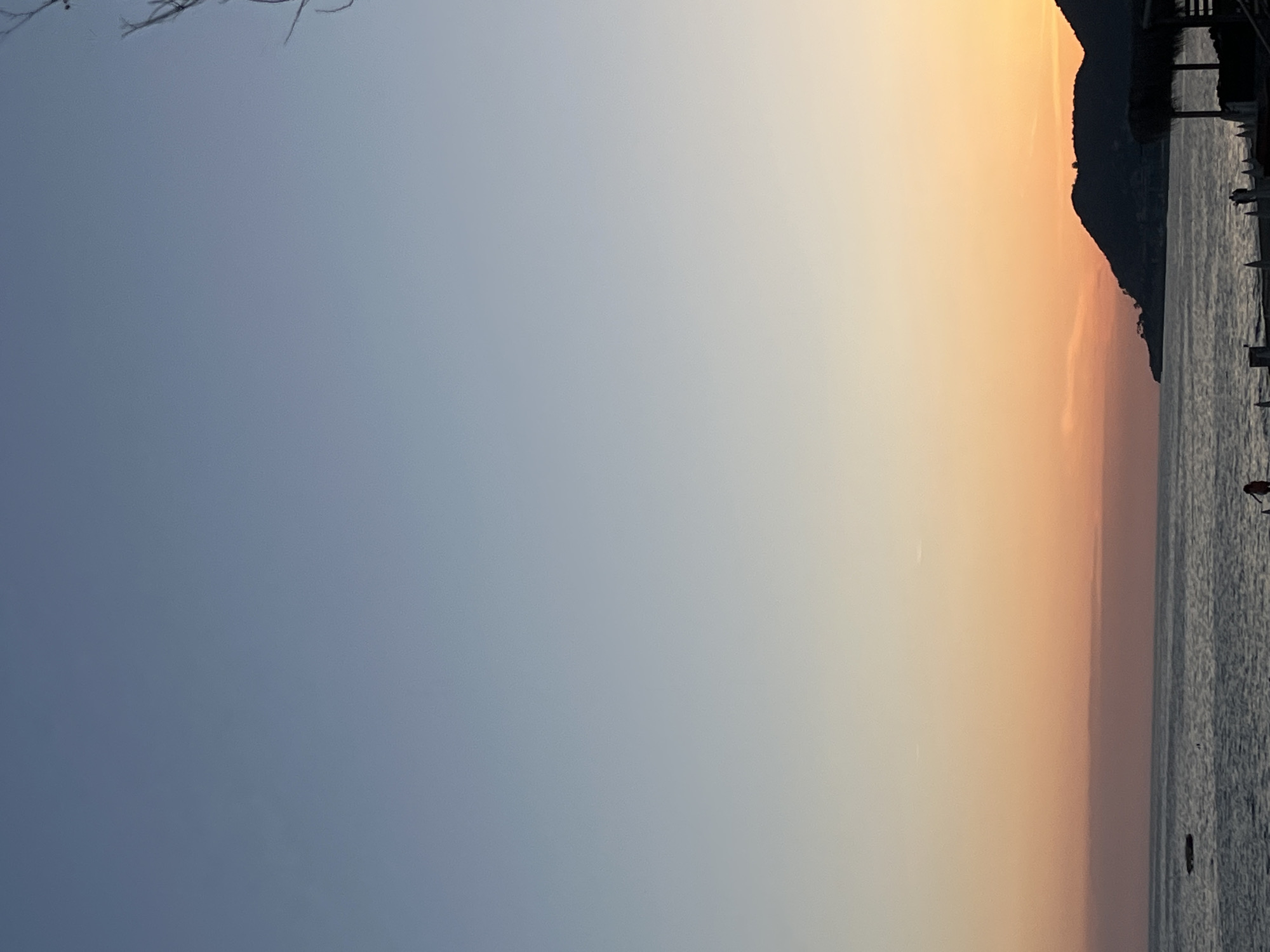The landscape image captures a serene coastal scene during either sunrise or sunset, dominated by a vast sky and calm water. The sky displays a clear gradient, transitioning from a soft, muted blue-grey on the left to warm, ethereal shades of pale orange and yellow towards the right, indicating the low angle of the sun. In the top-left corner, faint, bare tree branches extend into the frame, providing a subtle natural foreground.
A distinct horizon line separates the expansive sky from a tranquil body of water, likely the sea, which gently reflects the subtle colors of the sky. On the far right side of the image, a dark, prominent silhouette of a mountain range or large landmass rises from the water, sharply outlined against the warmer sky hues behind it. Closer to the bottom-right edge, along the water, vague dark shapes suggest boats or small structures, adding a hint of human presence to the otherwise natural scene. A small, reddish-orange object is faintly visible in the water near the extreme bottom right. The overall atmosphere is peaceful and calm, capturing the beauty of a coastal morning or evening. No visible text is present. This scene aligns with the geography of Alanya, Turkey, known for its coastal mountains and sea views.