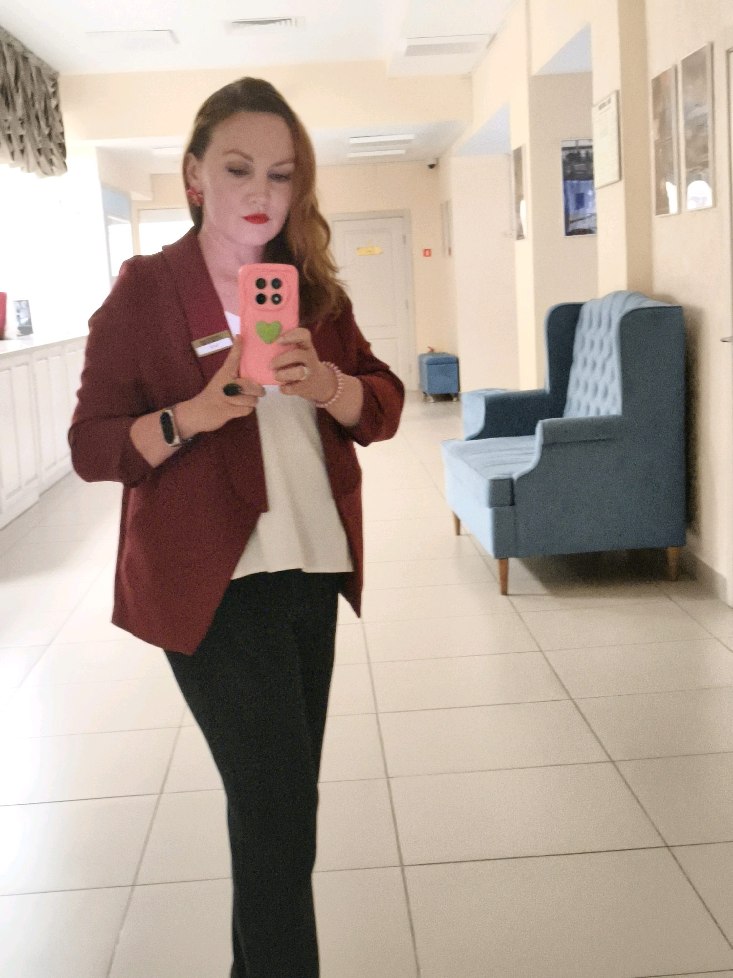A woman in a red blazer and black pants is taking a selfie in a well-lit corridor with light beige walls and floor tiles. She has auburn hair, bright red lipstick, and is wearing large red earrings and a pink phone case. A blue armchair is visible to her right, and a small blue ottoman is further down the corridor. The environment appears to be an indoor space, possibly a hotel or office building.