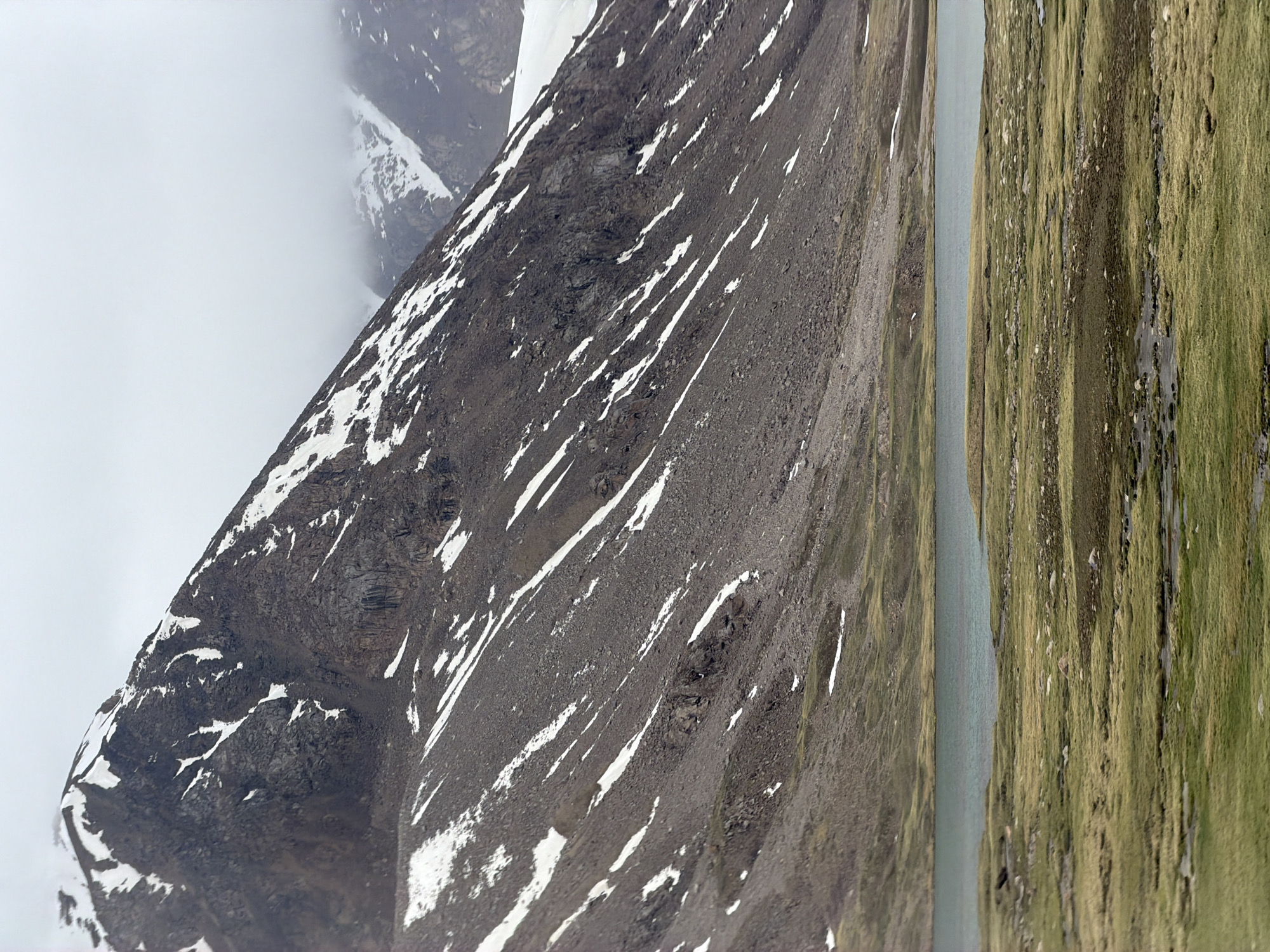 A vertical, medium shot captures a vast, mountainous landscape. A large, imposing mountain dominates the upper portion of the frame, its dark, rocky slopes streaked with patches of white snow. The summit is obscured by thick, white clouds. Below the mountain, a calm body of water stretches across the mid-ground, its surface a muted blue-green. The foreground features a grassy plain, dotted with scattered rocks and small pools of water, indicating a marshy or boggy terrain. The overall atmosphere is one of rugged natural beauty, with a sense of remoteness and cool, overcast weather.