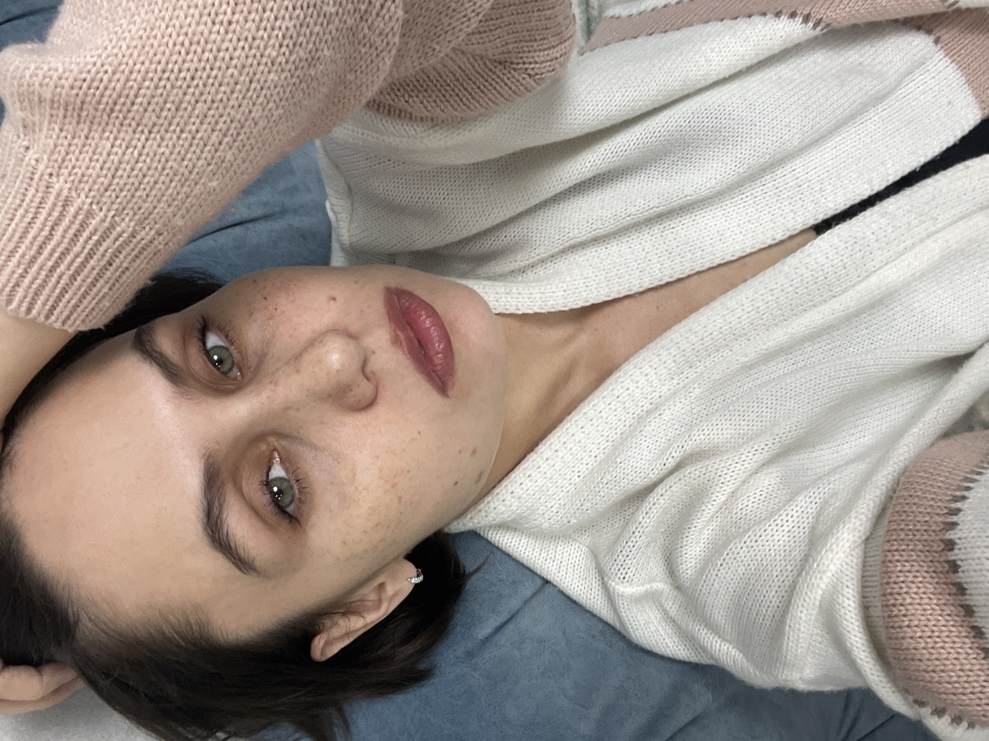 A woman with freckles is lying on a blue surface, possibly a bed or couch, wearing a white and pink knit sweater. She is looking directly at the camera with green eyes. Her hair is dark. She is wearing an earring in her left ear. The background is out of focus. There is no information to indicate that this image was taken in Izhevsk, Russia.