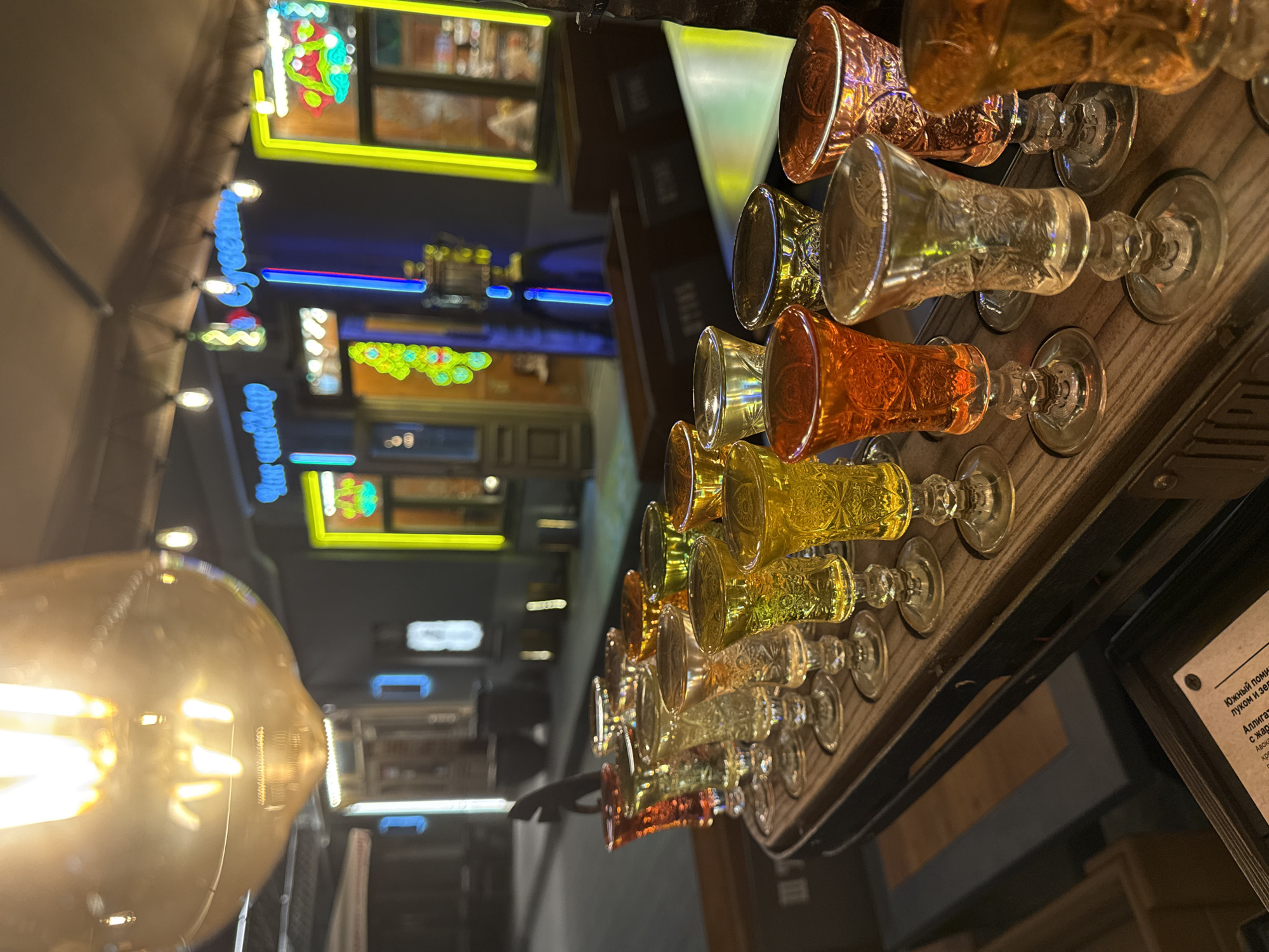The image shows a row of stemmed, patterned shot glasses filled with various colored liquids. The glasses are sitting on a wooden tray. In the foreground, a large, glowing light bulb is visible. The background reveals a blurred cityscape with neon lights of various colors and shapes. Based on the text visible in the image, this scene likely takes place in Kazan, Russia.