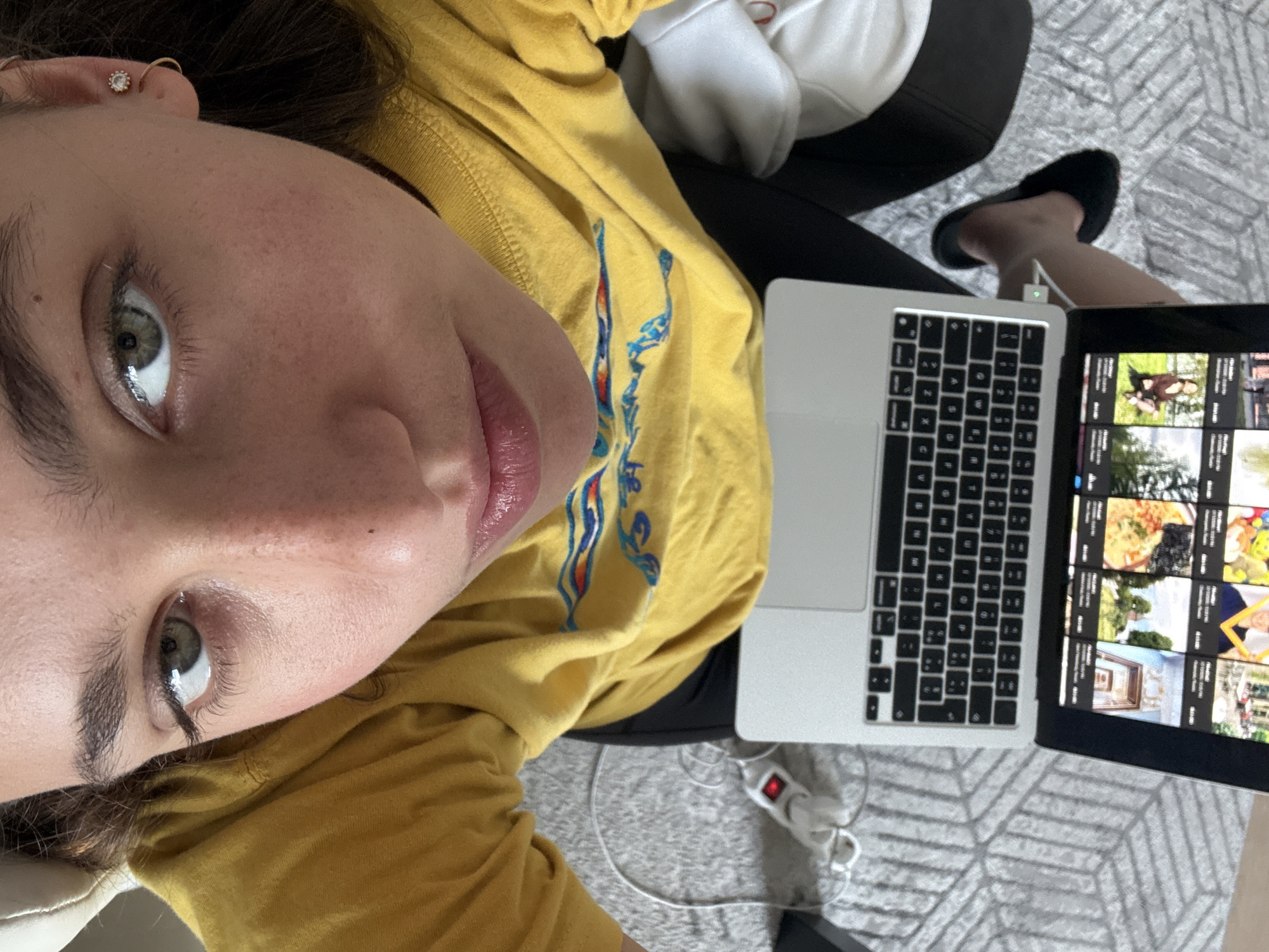 A person is seen in a close-up, top-down, selfie-style image, lying on a couch. The person has fair skin, hazel eyes, and light brown hair. They are wearing a yellow long-sleeved shirt with blue and red design on the upper left part of the chest. A laptop rests open on the couch with the screen displaying a grid of video thumbnails, suggesting the person is possibly browsing videos. A multi-outlet power strip sits beneath the laptop with some plugs visible. The person is wearing black pants and black slippers are visible on one foot. The couch is positioned on a gray carpet with a geometric pattern. The location in Milan, Italy is unknown.