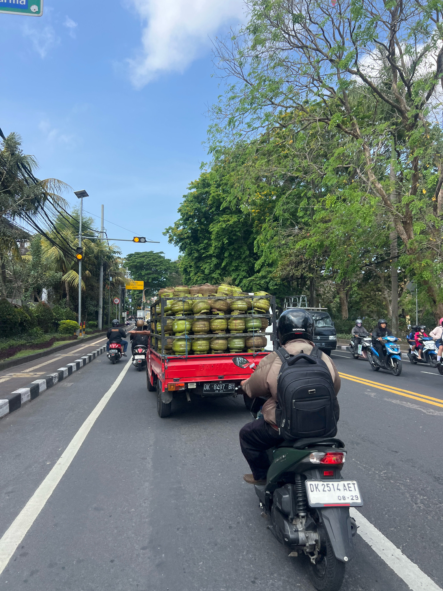 The image depicts a street scene in Denpasar, Indonesia, characterized by moderate traffic and lush greenery. In the foreground, a person wearing a brown jacket and a black backpack rides a dark green scooter. The scooter's license plate reads "DK 2514 AET" and "08-29". To the left, a red truck loaded with numerous green gas cylinders is a prominent subject. The truck's license plate is partially visible, showing "DK 8497 BH" and "08-25".
The road is paved with asphalt and marked with white and yellow lines, indicating traffic lanes. Several other motorcycles are visible in the background, navigating the road. On the left side of the street, there is a sidewalk with black and white striped borders, lined with manicured bushes and trees. Traffic signals with yellow lights are visible overhead, along with streetlights and a dense canopy of green trees that line the street. The sky is partly cloudy, with patches of blue. A yellow street sign is also present, though its text is not fully legible. The overall atmosphere suggests a typical day in a bustling urban environment with a strong presence of tropical vegetation.
