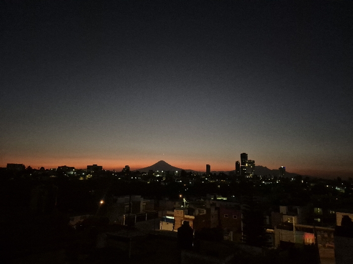 An elevated, wide-angle view captures a twilight or pre-dawn urban landscape in Puebla, Mexico. The sky occupies approximately two-thirds of the frame, exhibiting a gradient from dark charcoal grey at the zenith to a vibrant orange-red horizontal band at the horizon line. Centrally positioned against this horizon is a large, dark, symmetrical conical mountain, crowned by a solitary bright white point of light near its summit. To the right of the mountain, a cluster of approximately five to seven multi-story modern buildings forms a dense, illuminated skyline, with numerous distinct light sources visible within their structures. The urban sprawl extends across the midground and foreground, characterized by dark building masses, some with faint internal lighting or visible streetlights. The immediate foreground is dominated by the dark, indistinct forms of rooftops and the upper portions of closer residential or commercial buildings. No individuals, vehicles, or specific interactions are discernible. The environment is static, characterized by artificial illumination against natural low light conditions.