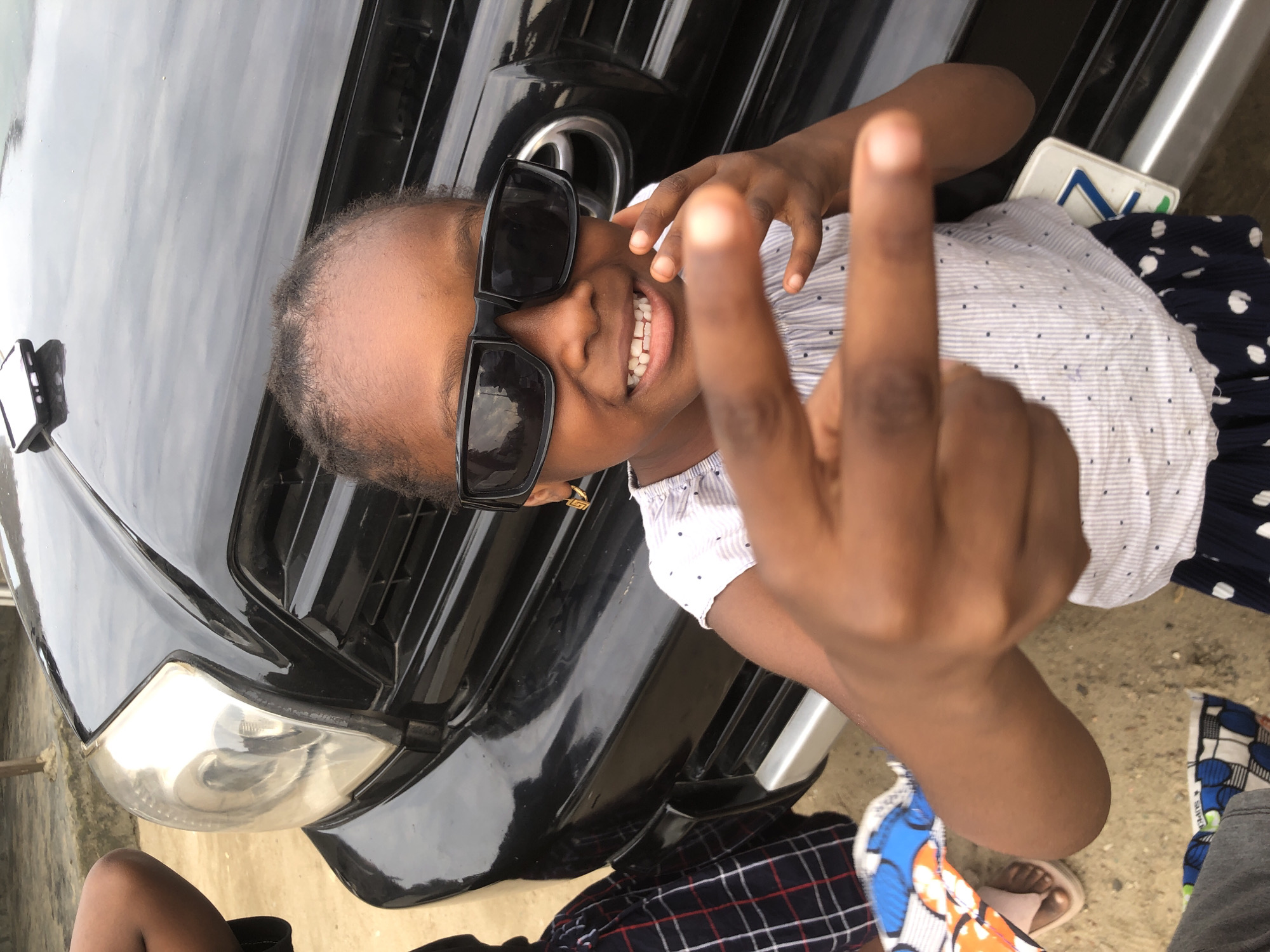 A young Black girl wearing sunglasses and a striped shirt stands in front of a black SUV. The SUV's hood and front grille are visible, as is its headlight. The girl is making a peace sign with her right hand, and her left hand is near her face. She is smiling and appears to be in a playful mood. The background is blurred, but it appears to be an outdoor setting, possibly a street or a parking lot. A phone is resting on the hood of the car. The image appears to have been taken in Nigeria, specifically in Ado.