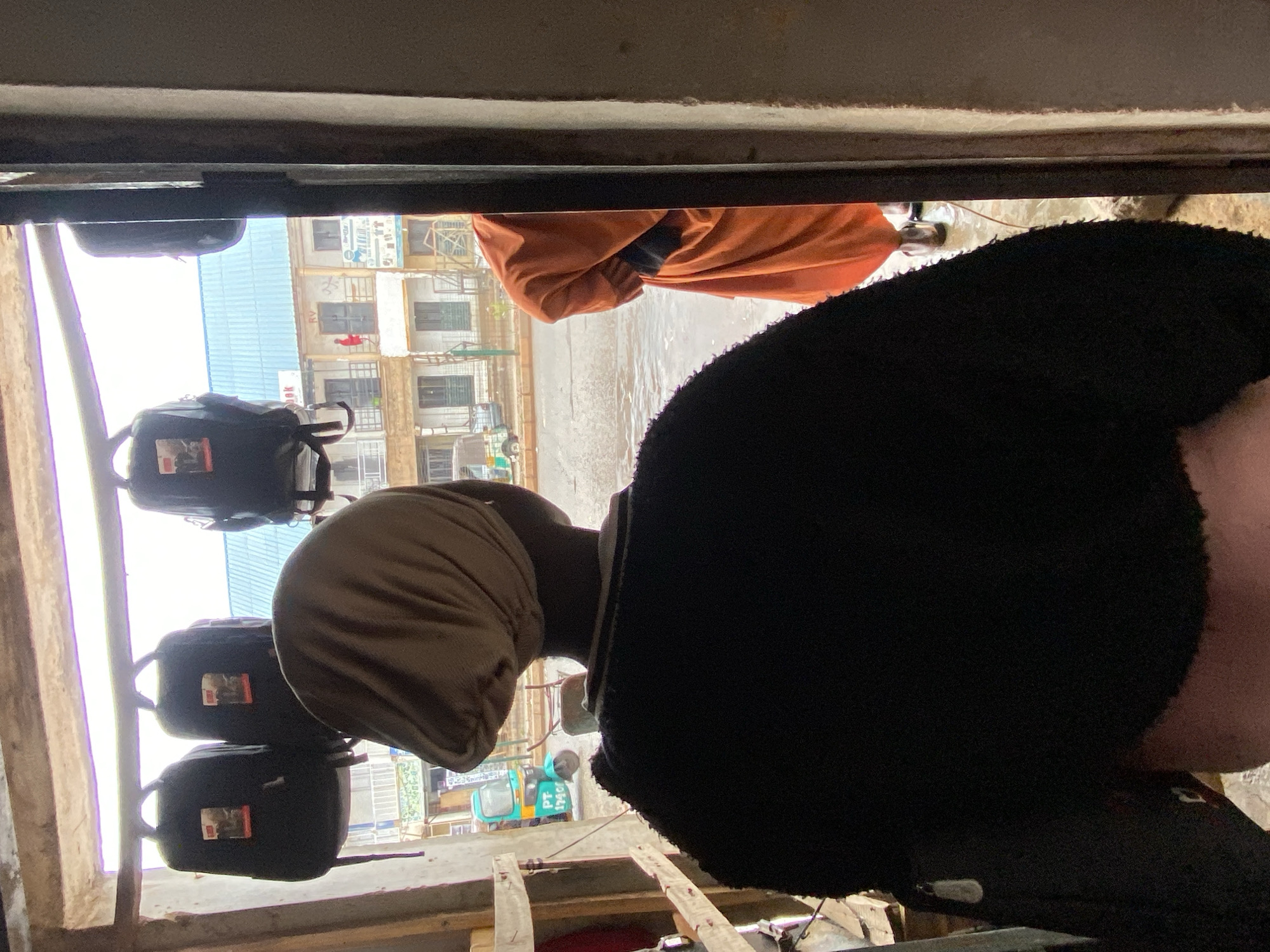 The image shows an eye-level view of a scene in Bukuru, Nigeria, possibly taken from inside a vehicle or building looking outwards. The foreground contains two people; one is wearing a black fluffy hat with dark clothing, and the other is wearing an orange shawl. The view through the opening reveals a street scene with a multi-story building, and a motorized rickshaw and pedestrians are also visible. There are also 3 backpacks hanging on what seems to be a bar.