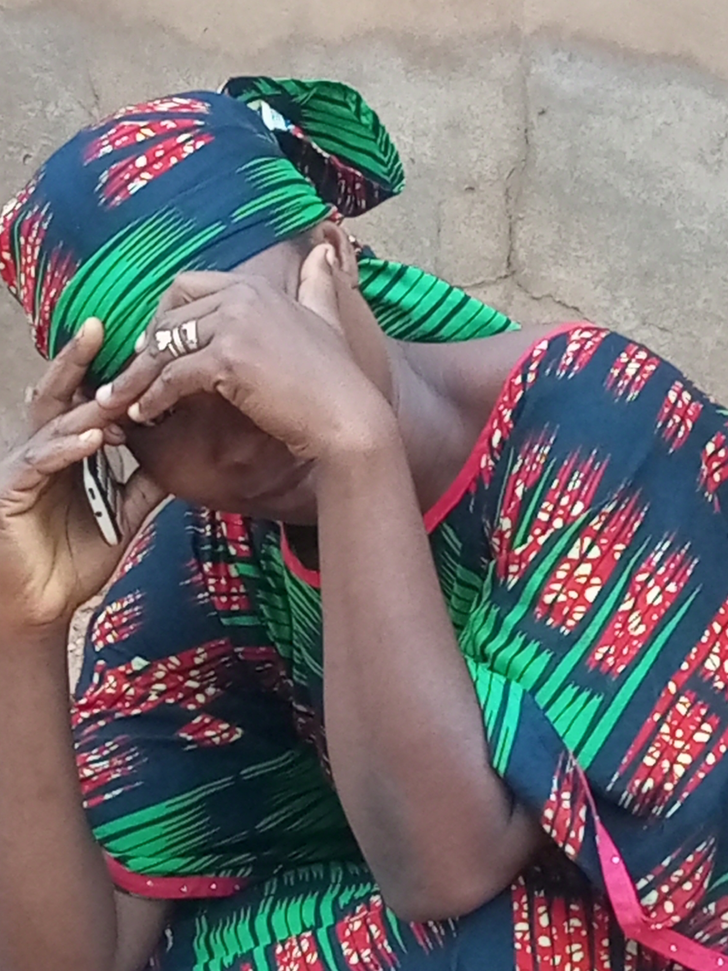 A close-up, medium shot shows a woman wearing a vibrantly patterned headscarf and dress. The fabric of both the headscarf and dress is dark blue with a repeating pattern of red and white abstract shapes, accented with bright green stripes and a pink trim on the dress. Her hands are raised to her face, covering her eyes. One hand, adorned with a silver-toned ring, is pressed against her forehead, while the other hand is positioned near her cheek. A portion of a white object, possibly a cell phone, is visible near her left ear. The background consists of a textured, beige wall that appears to be made of rough plaster or earth. The scene is set outdoors, as suggested by the natural lighting and the unfinished appearance of the wall. The woman's posture and the way she holds her hands suggest she may be experiencing distress or deep thought. The location is identified as Tempure, Nigeria.